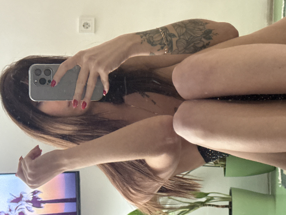A woman is taking a selfie in a mirror, holding her phone in her right hand, obscuring her face. She has long brown hair. A tattoo is visible on her right arm. She is wearing a black top with a sparkly pattern. A television screen is visible in the mirror’s reflection along with green plants in pots. A power socket is visible on the white wall. The image is rotated counter-clockwise.