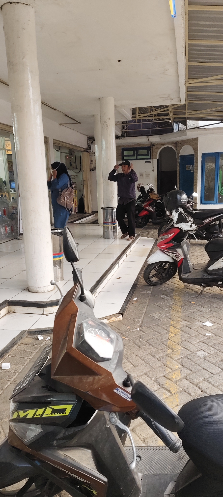 This image captures an outdoor scene in Malang, Indonesia, possibly in front of a building with white pillars and a tiled walkway. In the foreground, the front of a brown and black motorcycle is visible. Further back, a woman wearing a blue hijab and a mask stands near a white pillar, holding a backpack. To her right, a man in a dark jacket and pants, wearing a baseball cap, is adjusting his cap with both hands, appearing to be in mid-action. Several motorcycles are parked behind them, and in the background, architectural elements suggest a covered outdoor area with entrances and windows. The ground is a mix of tiles and paving stones.