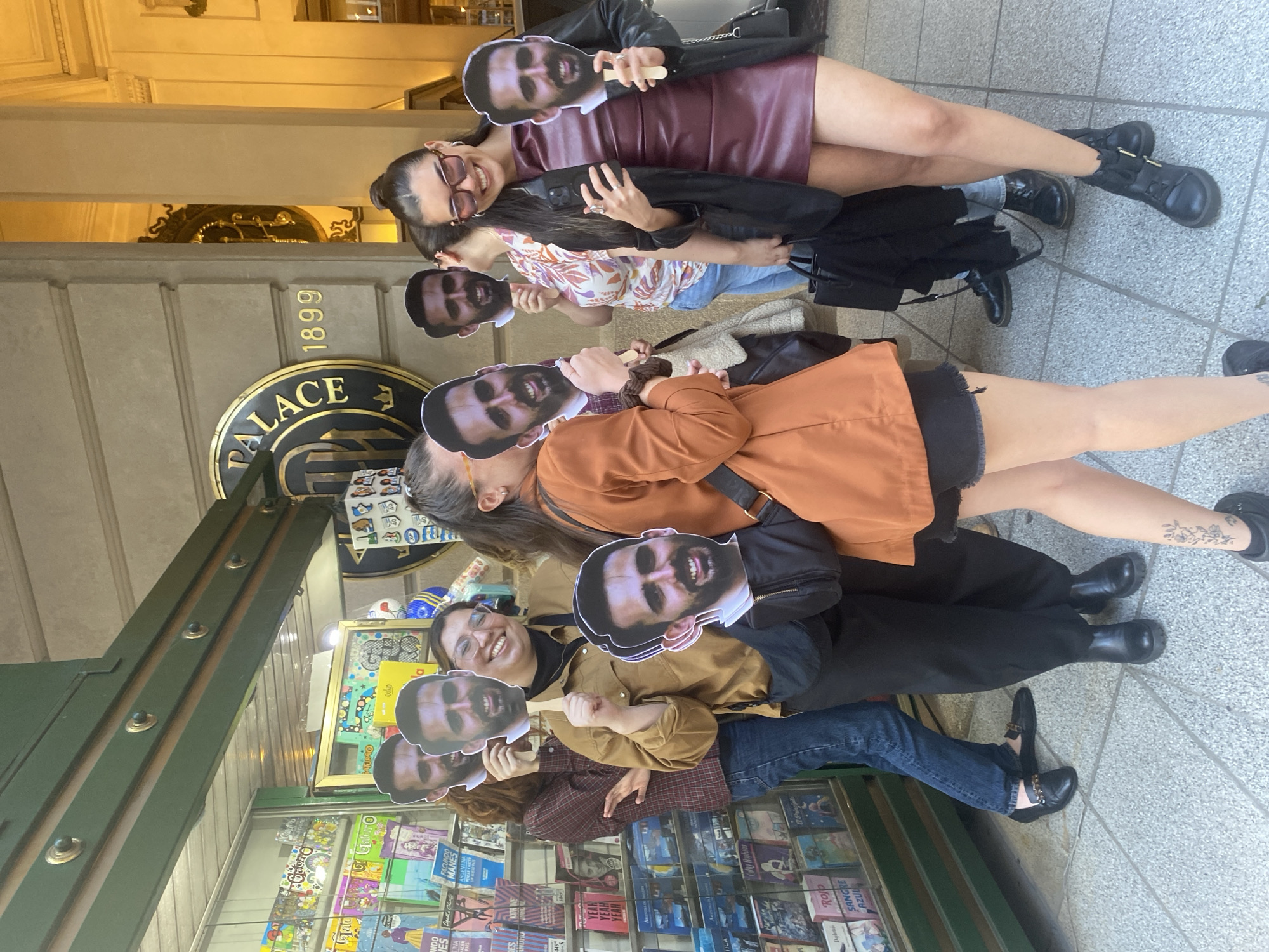 A group of five people pose for a photo in front of a newsstand in Buenos Aires, Argentina. The building behind them features the word "PALACE" and the date "1899" above the entrance. Each person holds up a printed headshot of the same man. The individuals, varying in age and attire, are standing closely together. Their clothing includes a mix of colors and styles, from casual jeans to more formal dresses. The newsstand displays a variety of magazines and books.