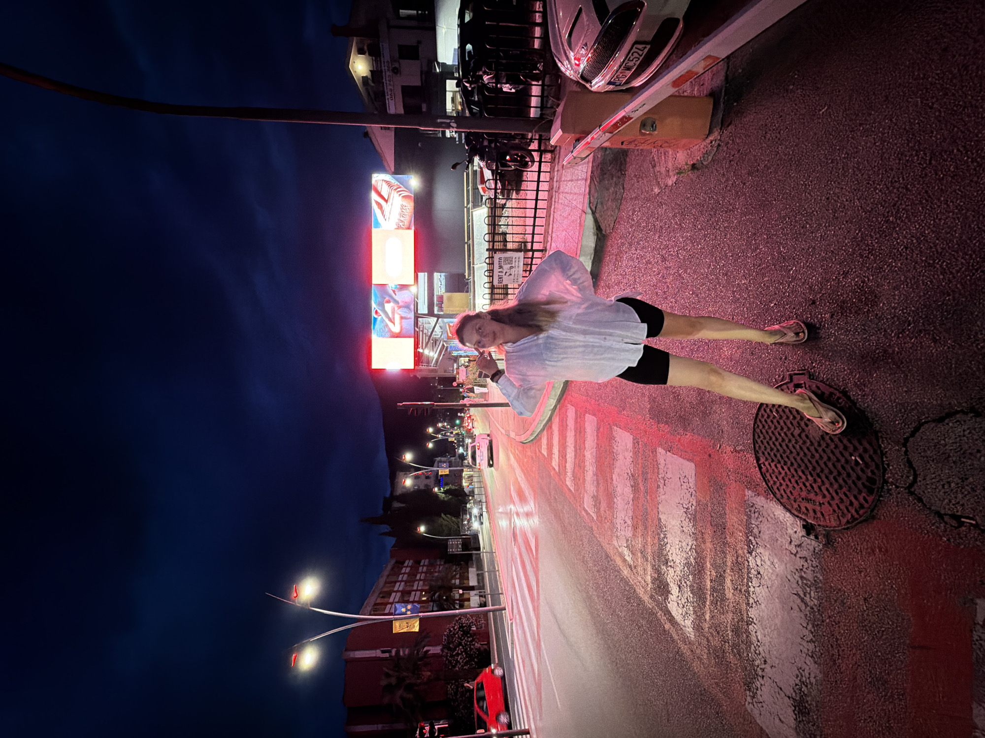 The image depicts a street scene at night in Budva, Montenegro. A young woman stands on a sidewalk near a crosswalk, illuminated by the red glow of streetlights and neon signs. She is wearing a white button-down shirt and black shorts, with her hand raised to her head. The street is wet, suggesting recent rain, and cars are visible driving in the background. A pedestrian crossing with white stripes is prominent in the foreground, reflecting the red light. In the background, there are buildings with illuminated windows, a Ferris wheel, and a billboard advertising "Rent a Moto." The sky is dark blue with some clouds, indicating it is nighttime. The overall impression is that of a lively urban environment with commercial activity.