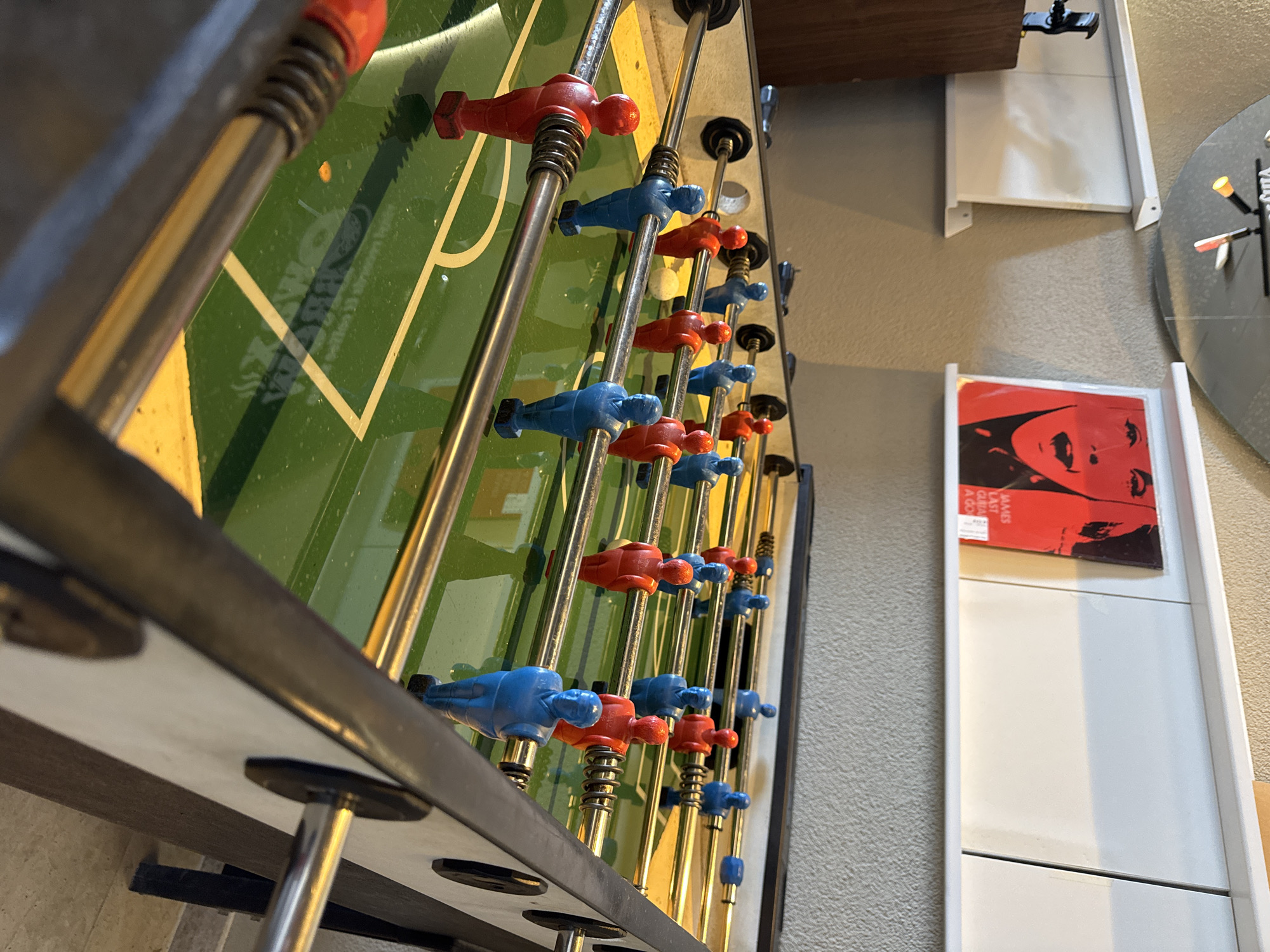 The image shows a foosball table with red and blue players, set against a wall with a beige texture. Behind the table, a white shelf unit is visible, holding a red album cover with a partial view of a face. Further in the background, parts of a gray decorative clock and a white wall-mounted shelf are seen. The scene is indoors, with the foosball table as the main subject. No people are visible. There is no information in the image to determine the location in the city of Rafailovići, Montenegro.