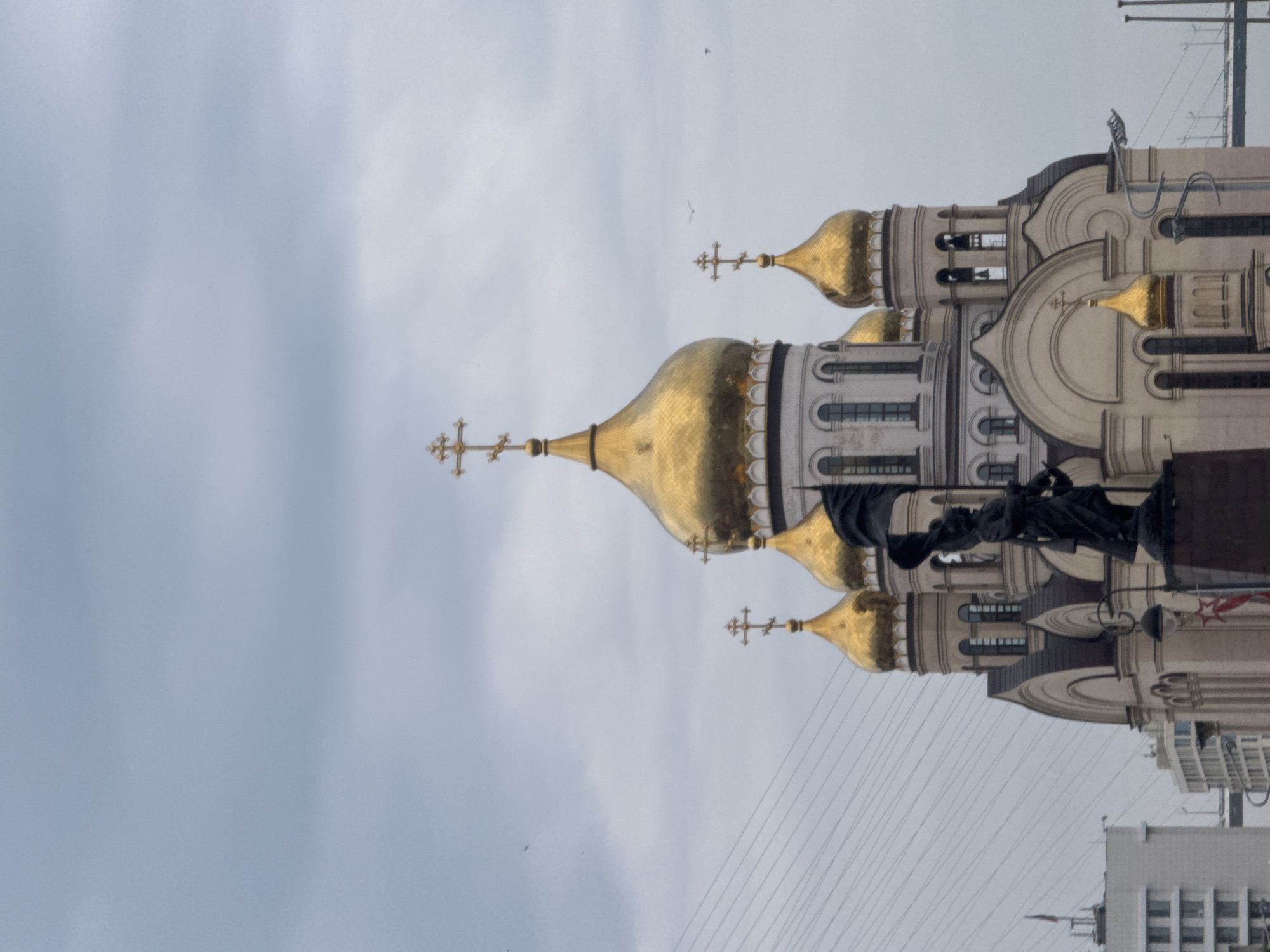 The image shows the Church of the Assumption of the Mother of God in Vladivostok, Russia. The church features several golden domes topped with crosses. In the foreground, a dark-colored statue of a person holding a flag is visible. The sky is overcast with clouds. In the background, a bridge and modern buildings can be seen.