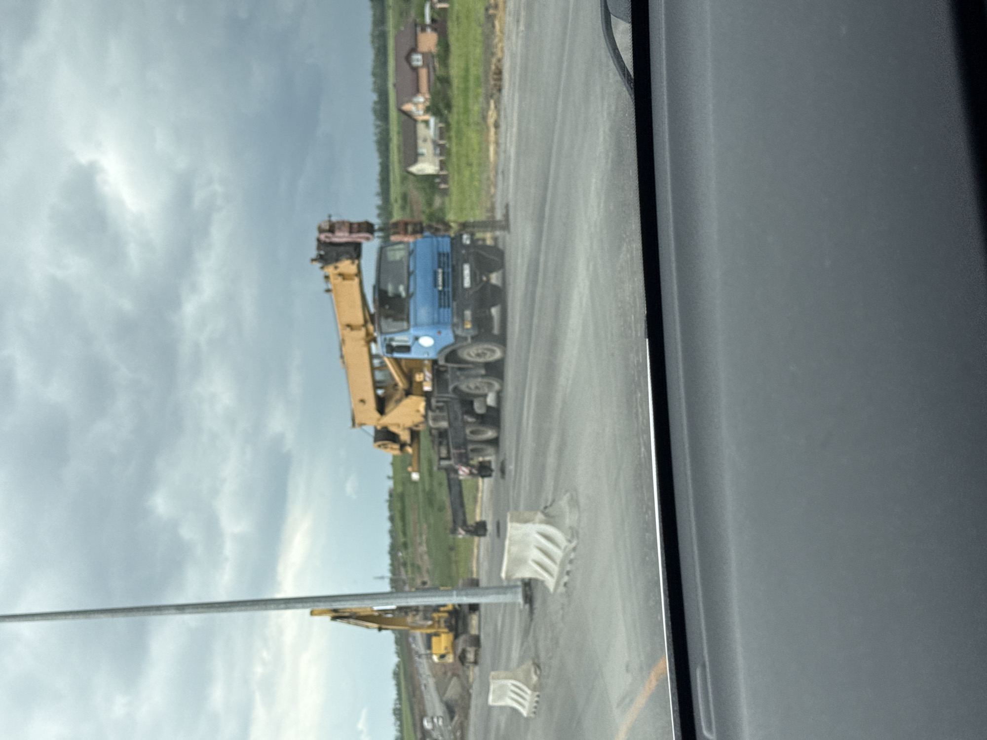 The image shows a scene in Shchepkin, Russia. A blue and black crane truck with a yellow crane arm is parked on the right side of a road. Adjacent to the crane are white and gray construction barriers. Further down the road, a yellow excavator is visible. The background includes a grassy field and houses. The image appears to be taken from inside a vehicle, with part of the vehicle's interior visible on the right side. The sky is cloudy.