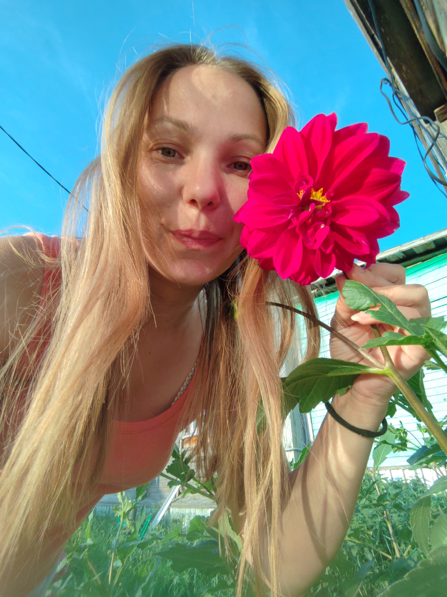 A woman with long blonde hair is holding a bright pink dahlia flower in front of her face. She is outdoors, likely in a garden, with a clear blue sky in the background. Green foliage and plants are visible in the foreground and background, suggesting a natural setting. A wooden structure, possibly part of a greenhouse or shed, is partially visible to the right. The image appears to be a selfie, with the woman looking directly at the camera. The location is specified as Chukhlem, Russia.