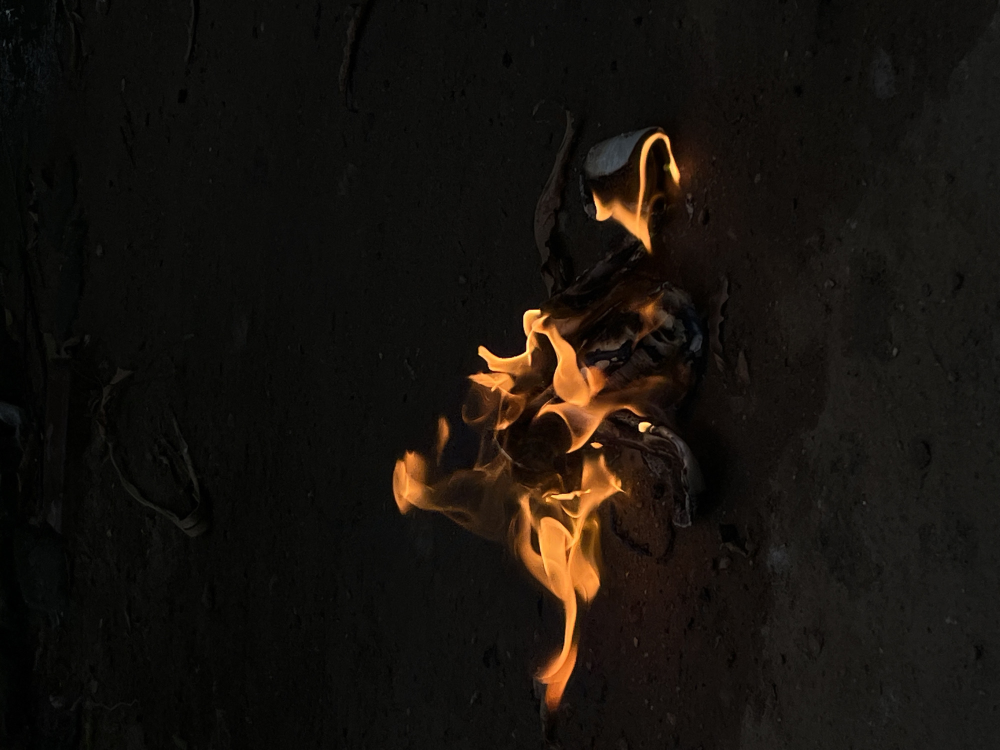 The image shows a small fire burning on the ground. The ground appears to be made of dirt or dark soil. The fire is small, with orange and yellow flames licking at the edges of what appears to be a burning piece of paper or fabric. The surrounding environment is dark, suggesting that the image was taken in low light. The location in Lokoja, Nigeria cannot be determined from this image.