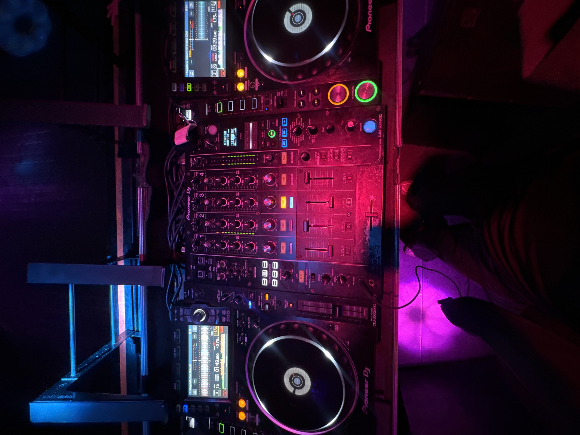 The image shows a Pioneer DJ setup consisting of two CDJ-2000NXS2 players and a DJM-900NXS2 mixer. The equipment appears to be set up in a DJ booth or performance area, likely within a nightclub or event venue. The lighting is a combination of red and blue, creating a vibrant and energetic atmosphere. Wires and cables are visible connecting the equipment.