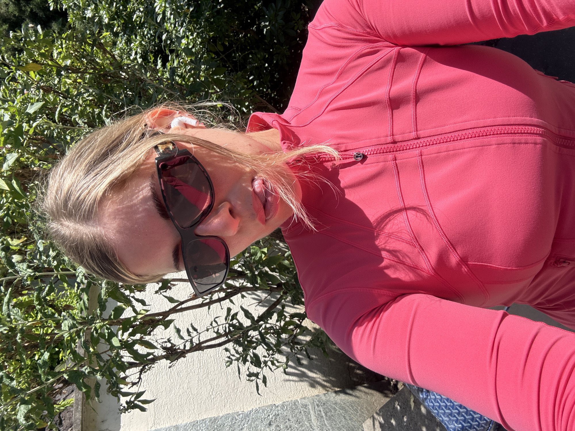 A blonde woman is taking a selfie outdoors. She wears sunglasses, earbuds, and a pink zip-up jacket. She is sticking her tongue out. The background features greenery and a light-colored wall. The setting could potentially be in Paris, France given the presence of classic architecture and greenery.