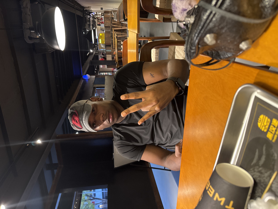 The image depicts an interior scene, presumed to be a restaurant or cafe, located in Liangjiang, China. A male subject with dark skin, wearing a grey baseball cap with a red 'B' logo and a black short-sleeved t-shirt, is seated at an orange wooden table, looking directly at the camera. The subject's right hand is raised, displaying three extended fingers (thumb, index, and middle), while the left arm rests on the table, revealing a small tattoo on the upper arm and a smartwatch on the wrist.
On the table in the immediate foreground, a black paper cup with yellow text is positioned on its side next to a rectangular metal tray containing a black paper liner with a yellow burger logo and Chinese characters translating to "Black Burger." To the right, a black leather bag adorned with silver studs and a purple fuzzy attachment rests on the table. The background shows a dimly lit interior with a dark ceiling, exposed beams, and multiple light fixtures, including a prominent black dome pendant light. Several empty wooden tables and chairs are visible, along with a window on the left displaying an indistinct exterior view.