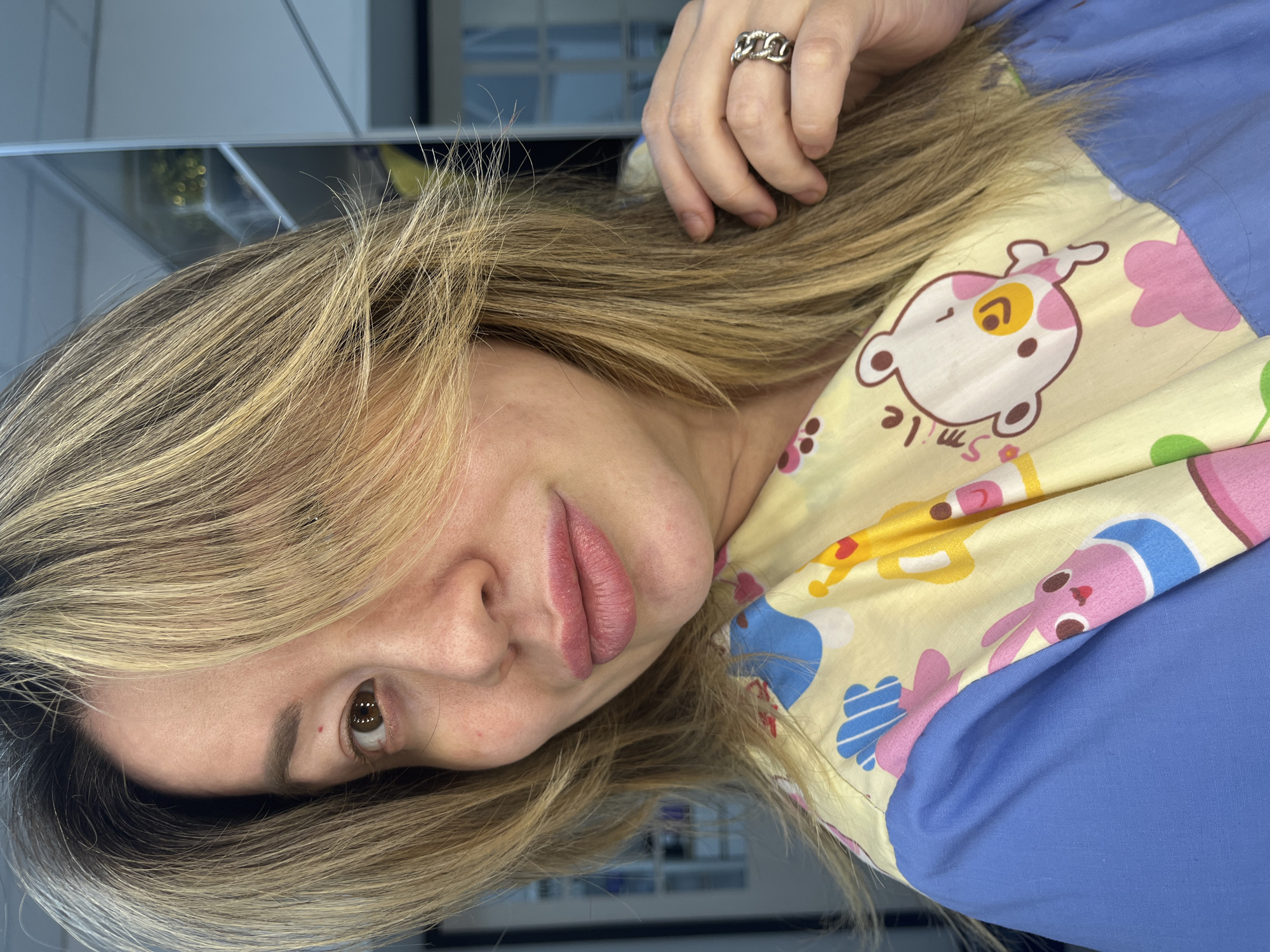 This rotated close-up portrait, captured in Perm, Russia, features an adult woman looking directly towards the viewer. The image is rotated 90 degrees counter-clockwise. She has long, straight blonde hair with darker roots, which falls across her face and shoulders. Her left eye is visible, a warm brown color, while her expression is neutral with slightly parted, natural pink lips.
The woman is wearing a blue top partially obscured by a bright yellow fabric panel. This panel is adorned with a playful pattern of cartoon characters, including a white dog-like figure with pink ears and a yellow snout, beneath which the word "smile" is printed upside down. Other elements on the yellow fabric include pink flowers and cartoon bunnies in blue and pink. On her left hand, a prominent silver ring with a chunky, chain-link design is visible, resting near her hair. The background is out of focus, appearing as a blurred reflection in a glossy surface, hinting at an indoor setting.