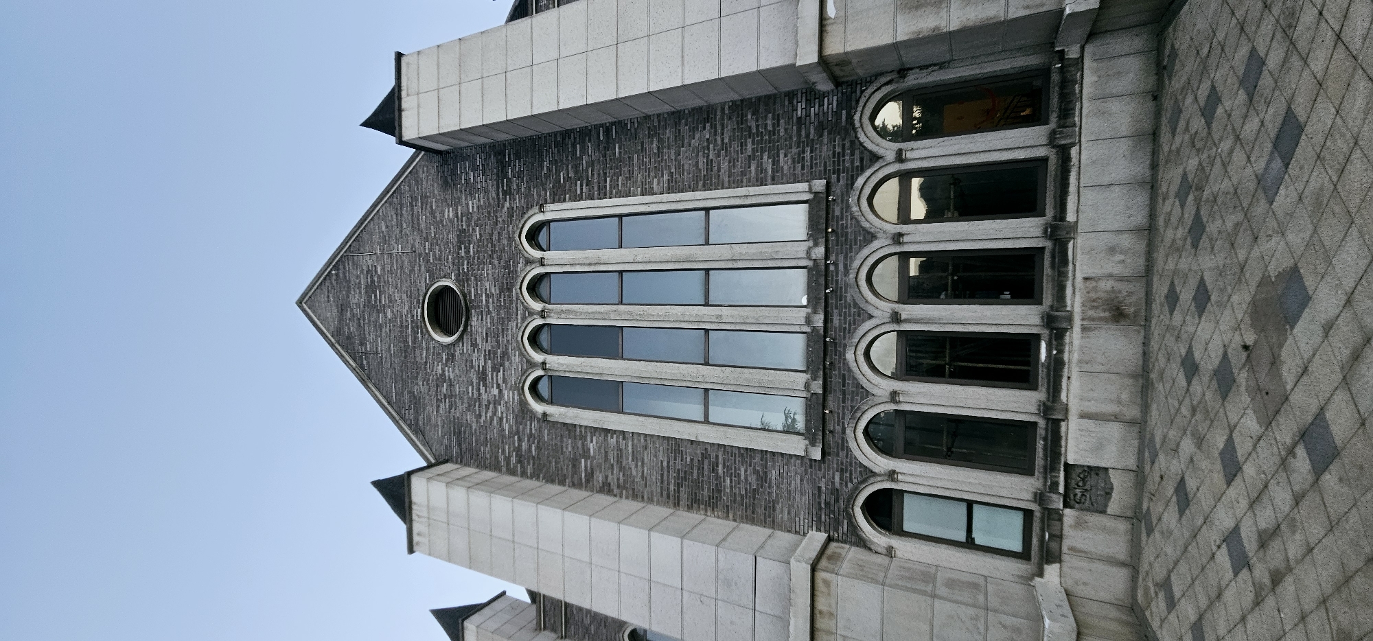 The image shows the facade of a building with a gothic architectural style. The building is constructed with dark gray bricks and accented with lighter stone elements. It features a prominent gable roof with two pointed spires on either side of the main structure. A row of tall, arched windows with thin mullions spans the upper portion of the facade, reflecting the clear, light blue sky. Below these, a second set of smaller, arched windows with dark panes is visible. The ground in front of the building is paved with a diamond pattern of light and dark gray tiles. There is a small, circular vent near the peak of the gable. The scene appears to be located in the Chongqing High-tech Zone, China, based on the provided context. No people or specific actions are depicted in the image.