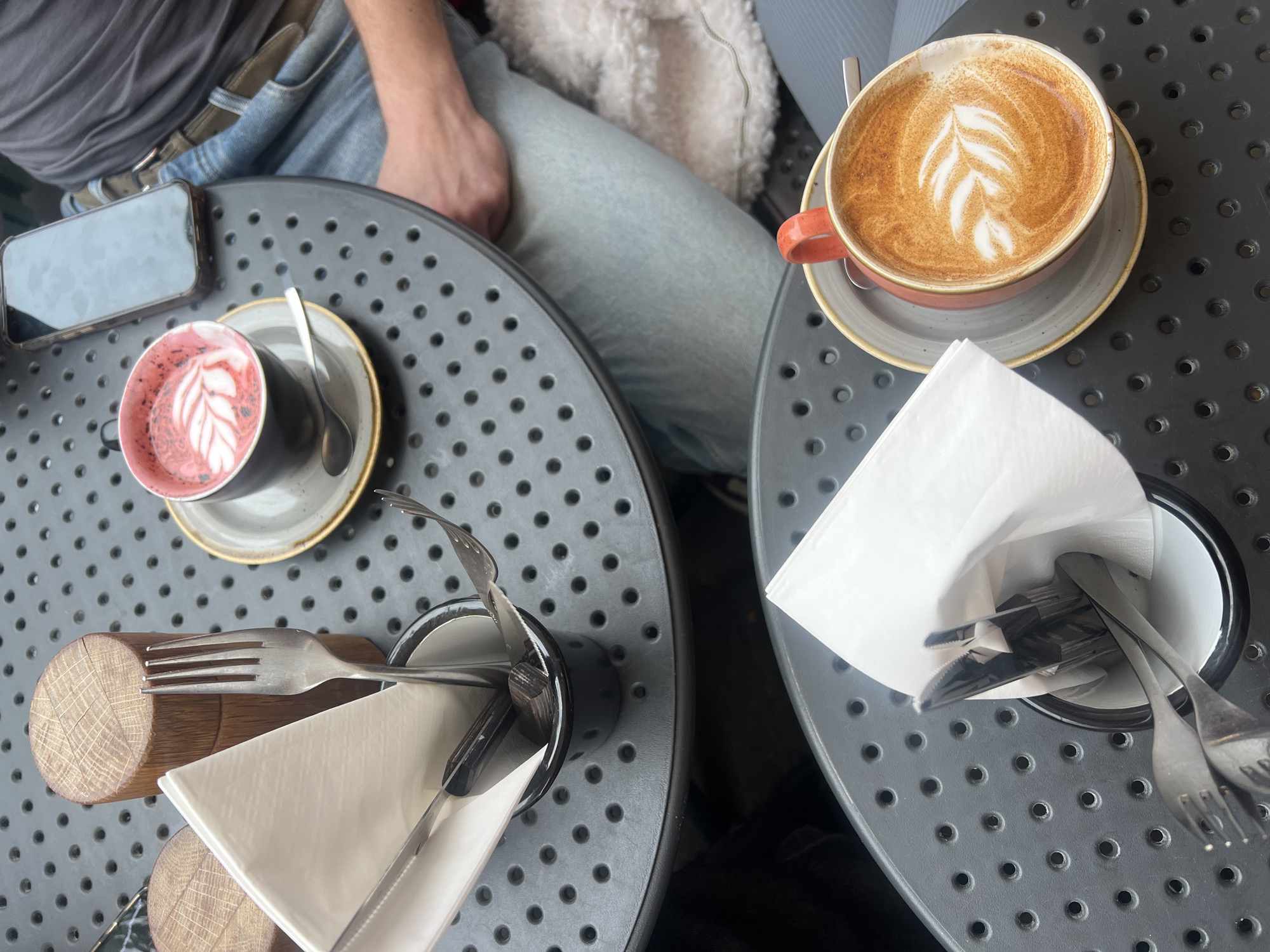 The image shows an overhead view of two circular tables on a gray, perforated surface, likely an outdoor cafe in London, United Kingdom. Two people are seated at the tables; one is wearing blue jeans and a gray top, and the other is wearing light-colored jeans and a furry white jacket. On the left table, there is a cell phone, a black cup containing a red beverage with latte art, a small spoon on a saucer, a wooden block supporting a metal cup filled with silverware and a white napkin. The right table holds a coral-colored cup with a cappuccino-style beverage and latte art on a saucer, and a metal cup filled with silverware and a white napkin. The scene appears to be a casual social gathering at a cafe.