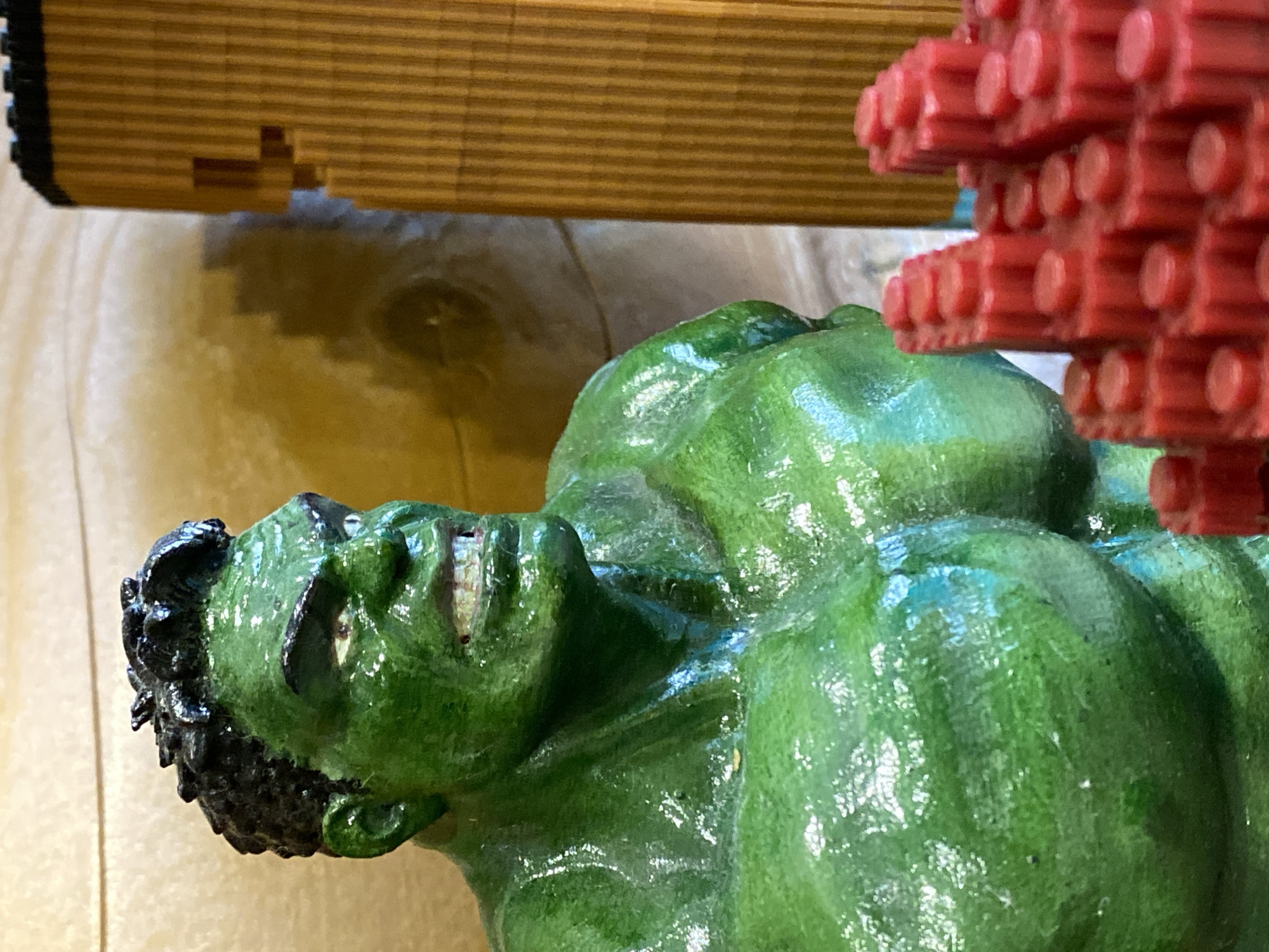 A close-up, angled photograph depicts a painted green Hulk figurine lying on a wooden surface. The figurine's head is oriented towards the left of the frame, with its face visible. Adjacent to the Hulk figurine is a red object composed of stacked, cylindrical elements, resembling building blocks. Above these objects, a strip of horizontal wooden slats is visible. There is no information about the location or the city of Chaikovsky, Russia, available from the image.