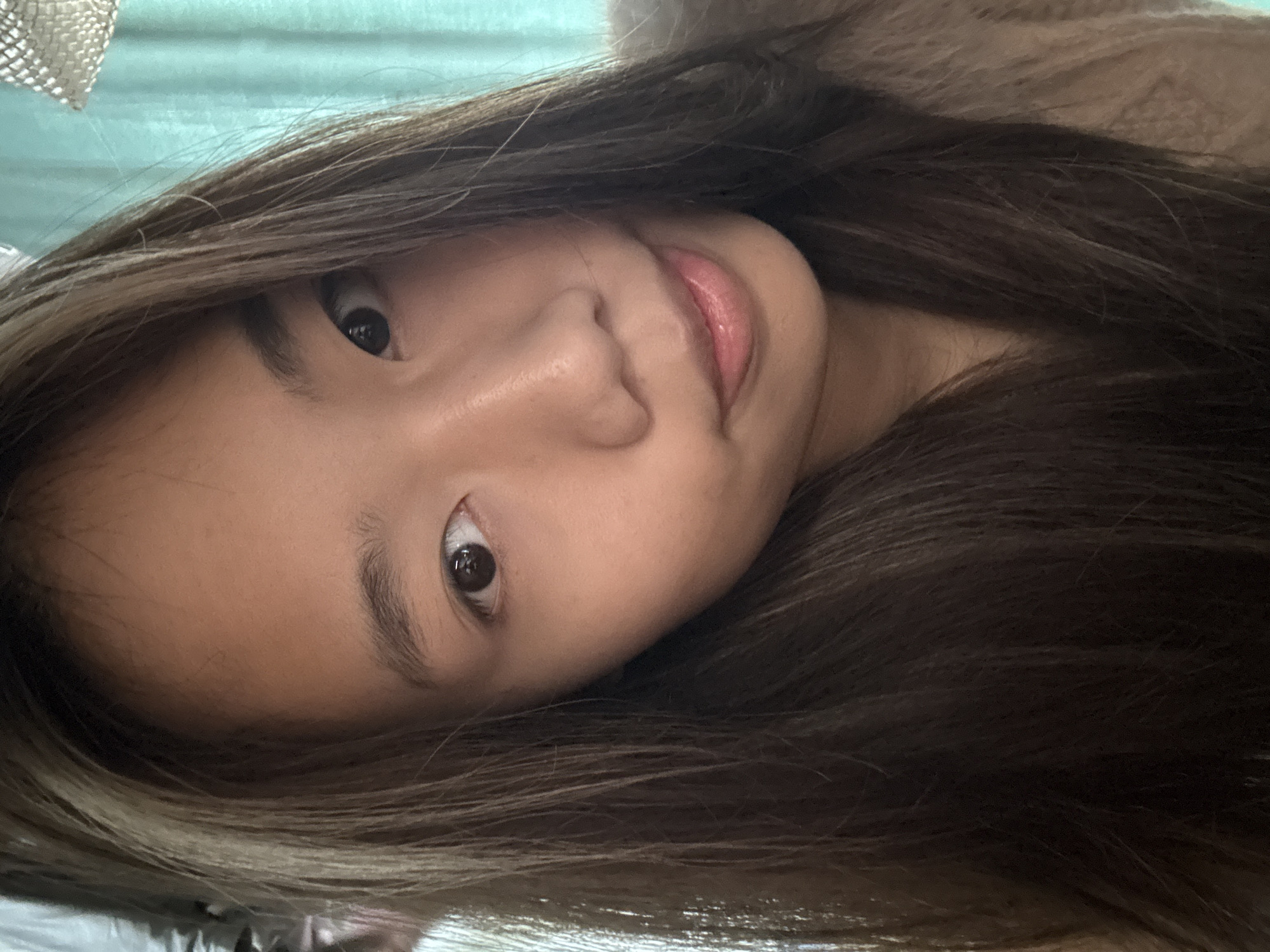 A close-up, slightly angled portrait of a young woman with long, dark brown hair framing her face. Her skin tone is fair, and her expression is neutral. The background appears to be a soft, light blue or green, possibly a curtain or wall. The location cannot be determined from the image, and there is no evidence to suggest the Nan'an District, China.