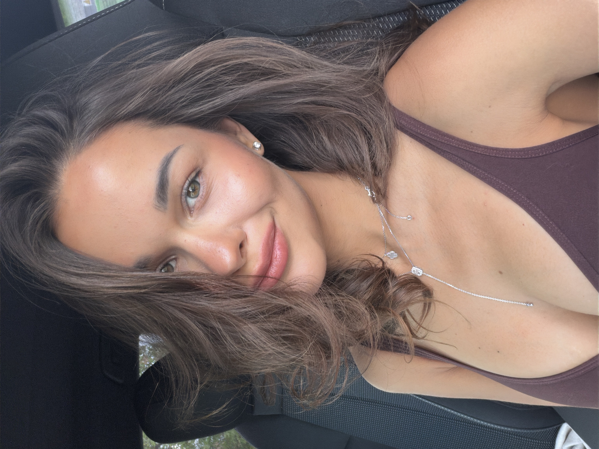 A young woman with fair skin and dark brown hair is taking a selfie inside a car. She's wearing a brown tank top and a silver necklace. Her hair partially covers her face, but her right eye is clearly visible. The background shows the car's interior. The location cannot be determined to be in Krasnoyarsk, Russia based on the available information.