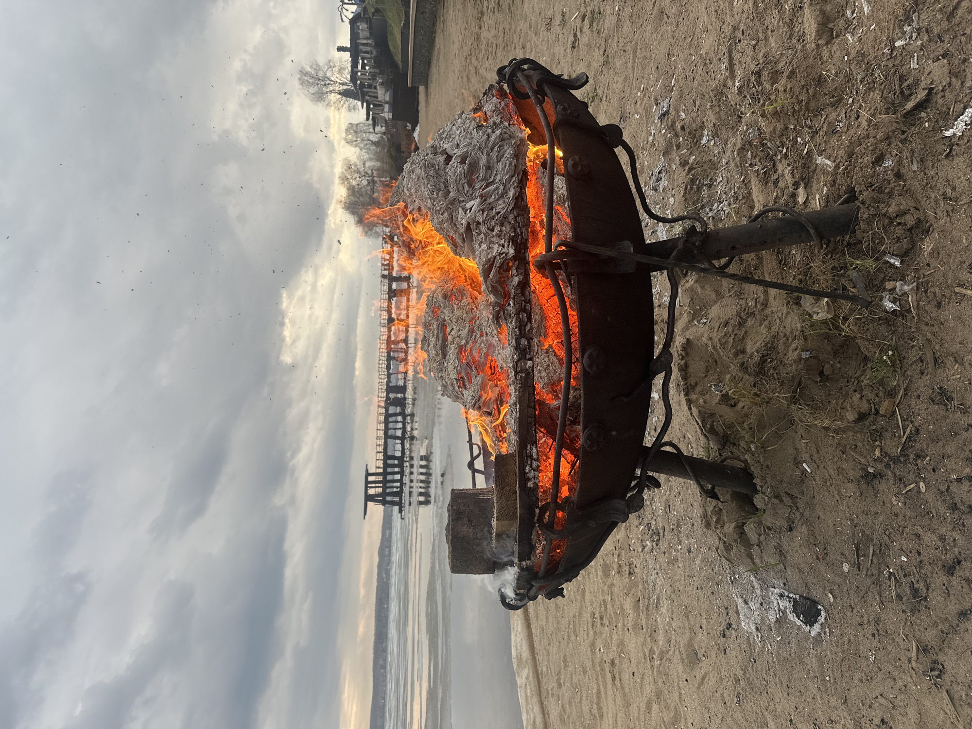 A metal fire pit stands on a sandy area, filled with burning material and flames. In the background, there is a body of water with a wooden pier extending into it. On the opposite shore, there is a building and trees. The sky is overcast with clouds. The location is likely near the Votkinsk Reservoir in Votkinsk, Russia.