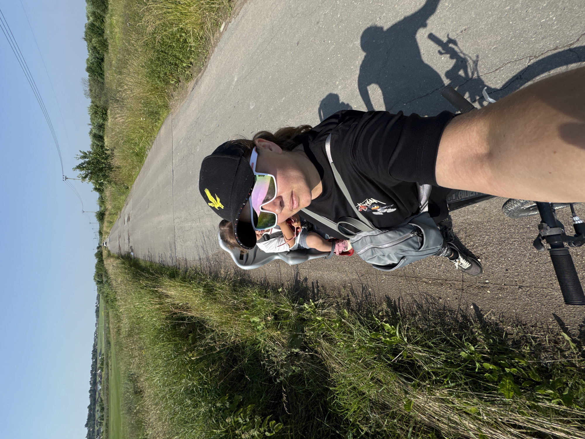 A woman wearing a black t-shirt, black baseball cap with a yellow eagle emblem, and white mirrored sunglasses is riding a bicycle on a paved road. She is taking a selfie, her right arm extended. Behind her, a child is seated in a child seat on the bicycle. The child is wearing a white t-shirt and gray shorts, with pink shoes. The road is bordered by tall, dry grass and a field on the left, and a line of trees on the right. In the distance, houses and power lines are visible under a clear blue sky. The scene appears to be in Kempelevo, Russia, on a sunny day.