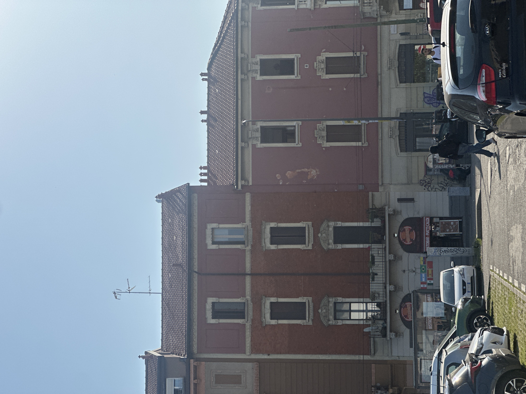 The image depicts a street scene in Milan, Italy, featuring a three-story building with a red facade and a tiled roof. The building has rectangular windows with decorative stone surrounds. At street level, there is a business, possibly a bar or bistro, indicated by signs.
Several parked cars line the right side of the image, occupying the street adjacent to a strip of green grass. Pedestrians are visible on the sidewalk, with one individual appearing to walk between parked cars. A black Kia car is parked on the right side of the frame.
An antenna extends from the roof of the building on the left.
