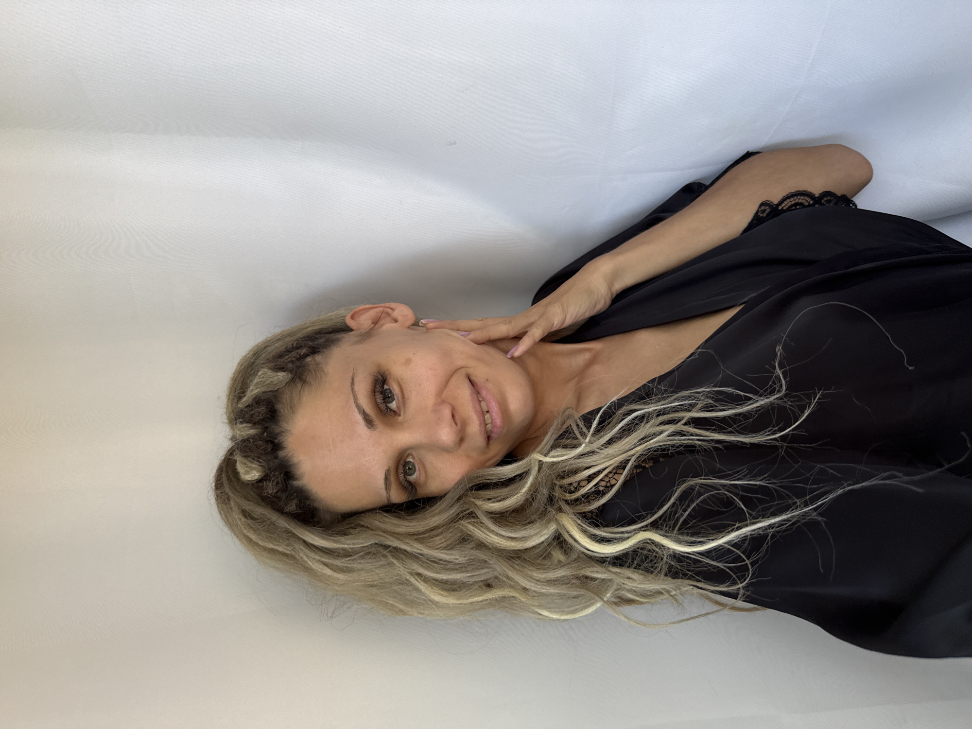 A light-skinned woman with blonde, curly hair is shown from the chest up. She is wearing a black robe and has her right hand resting on her neck. The woman is facing the camera and smiling. The background is a white curtain. The image is well-lit and in focus. There is no identifiable information in the image to suggest it is located in Rusanovo, Russia.