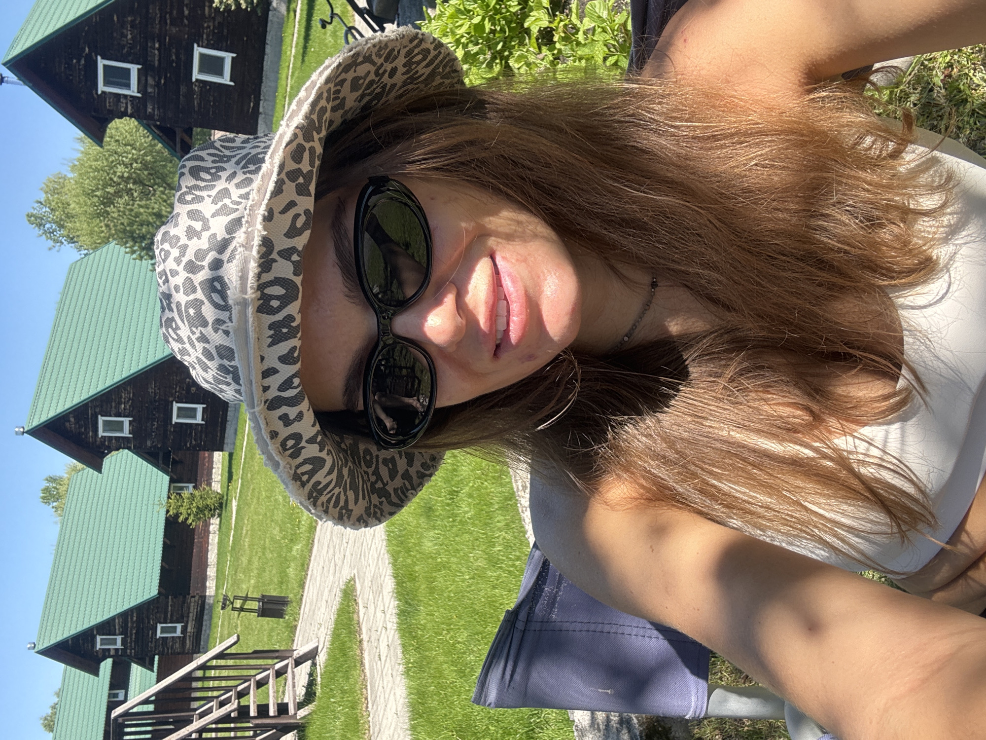 A young woman with long, light brown hair and sunglasses poses for a selfie. She is wearing a white top and a spotted bucket hat. In the background are several identical wooden cabins with green roofs, set amidst a grassy lawn and paved walkways. This scene is in Aya, Russia, as indicated by the location metadata.