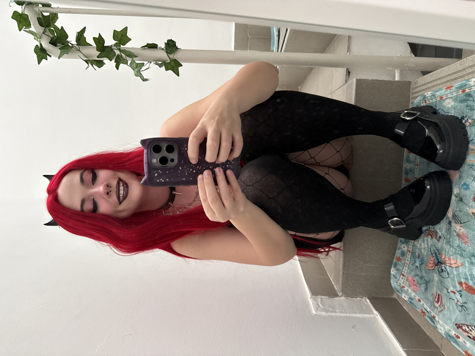 A person with red hair and small black horns poses in front of a mirror, holding a phone with a patterned case. The individual wears dark lipstick and a spiked collar. They are dressed in black fishnet tights and patterned thigh-highs. Black, shiny platform shoes are on their feet. A vine decoration hangs on the white railing above. A tiled staircase is in the background, and a patterned rug sits on one of the steps.