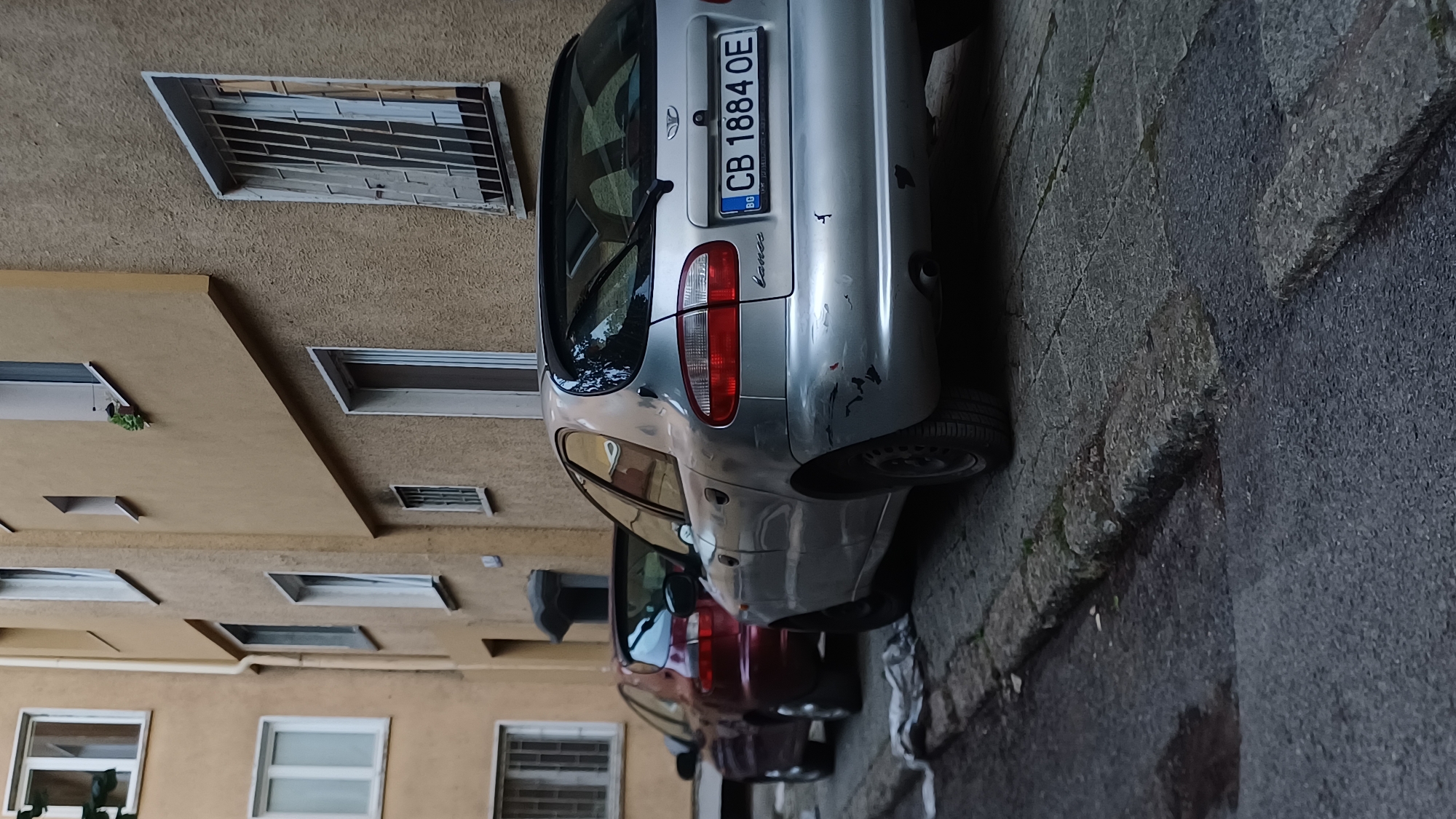 A low-angle, medium shot captures the rear of two parked cars on a street in an urban environment. The car in the foreground is a silver hatchback with minor damage to its rear bumper, featuring a license plate that reads "CB 1884 OE" with a "BG" country code. To its left and slightly behind is a dark red hatchback. Both cars are parked parallel to a weathered concrete curb. The background consists of a building with textured, light brown walls, featuring several windows. One window on the right is covered by a metal grate. The scene suggests a residential area or a street with limited parking.