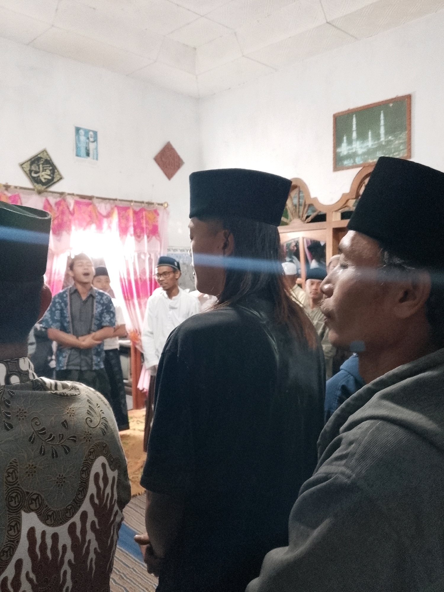 The image depicts a gathering of men indoors, likely in a home or community space in Rejoyoso, Indonesia. The scene is brightly lit, with a focus on the individuals present. Several men are wearing traditional Islamic attire, including black peci hats.
In the foreground, a man in a black peci is partially visible from the side, looking towards the center of the room. To his left, another man in a black peci with long hair is also seen from the side, facing away from the camera.
Further back, a group of men are engaged in conversation or prayer. One man in a patterned blue and grey shirt is speaking with his mouth open, possibly mid-sentence. Another man in a white outfit and glasses stands to his right, looking towards the speaker. Other individuals are visible in the background, partially obscured.
The walls of the room are white, adorned with decorative items. A framed image of a mosque with minarets hangs on the right wall, while a diamond-shaped calligraphy artwork and a framed picture of two figures are visible on the left wall. Pink and white patterned curtains hang across a window or doorway in the background, allowing light to filter through. The floor appears to be covered with a woven mat and a striped rug. The overall atmosphere suggests a religious or social gathering.