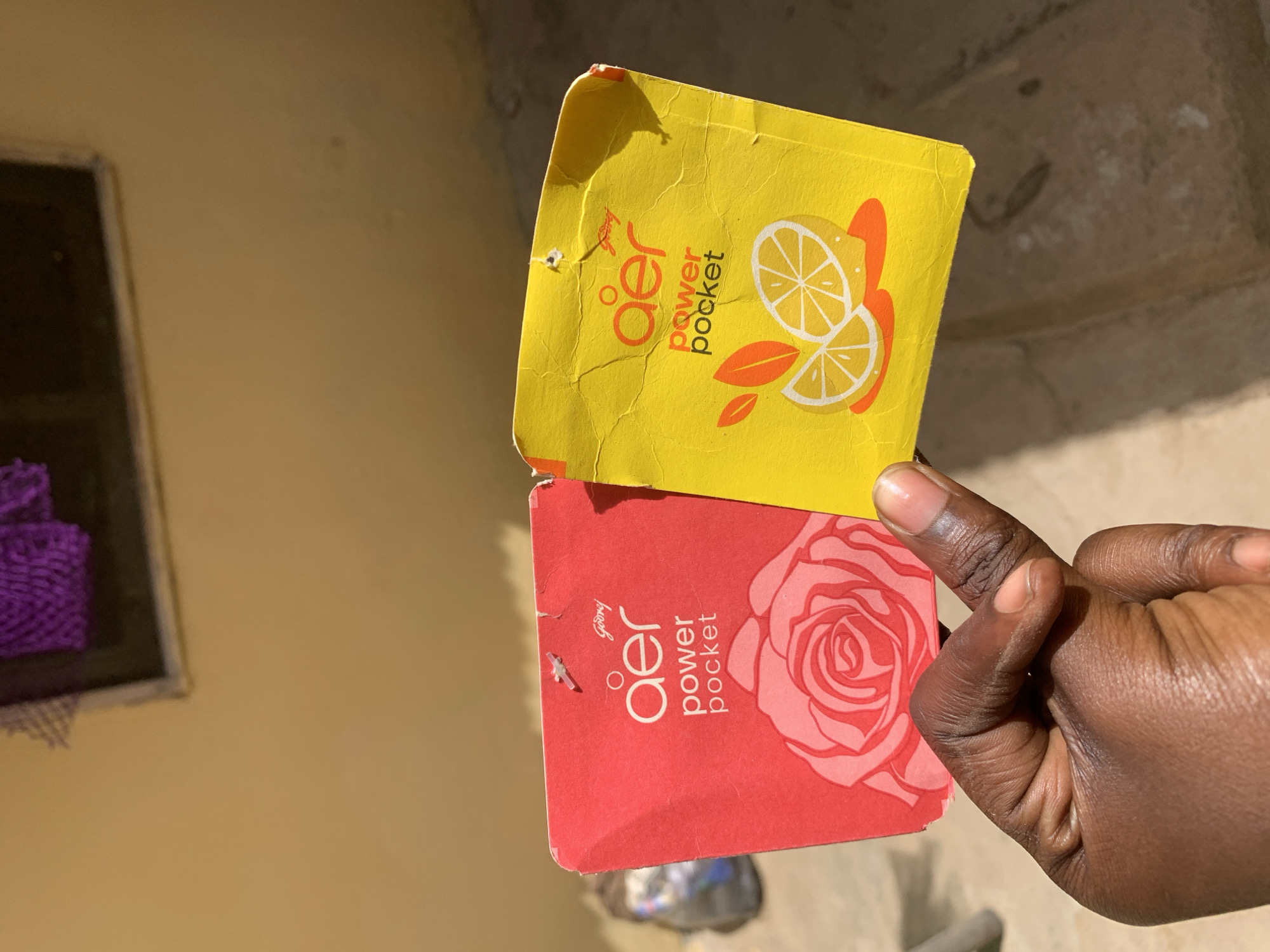 A medium shot shows a right hand holding two "aer power pocket" air freshener packages. The top package is yellow with a lemon graphic, and the bottom package is red with a rose graphic. The fingernail on the index finger of the hand is visible. A window with a purple object inside is visible in the background, along with a tan-colored wall. The location is Rafin Par, Nigeria.