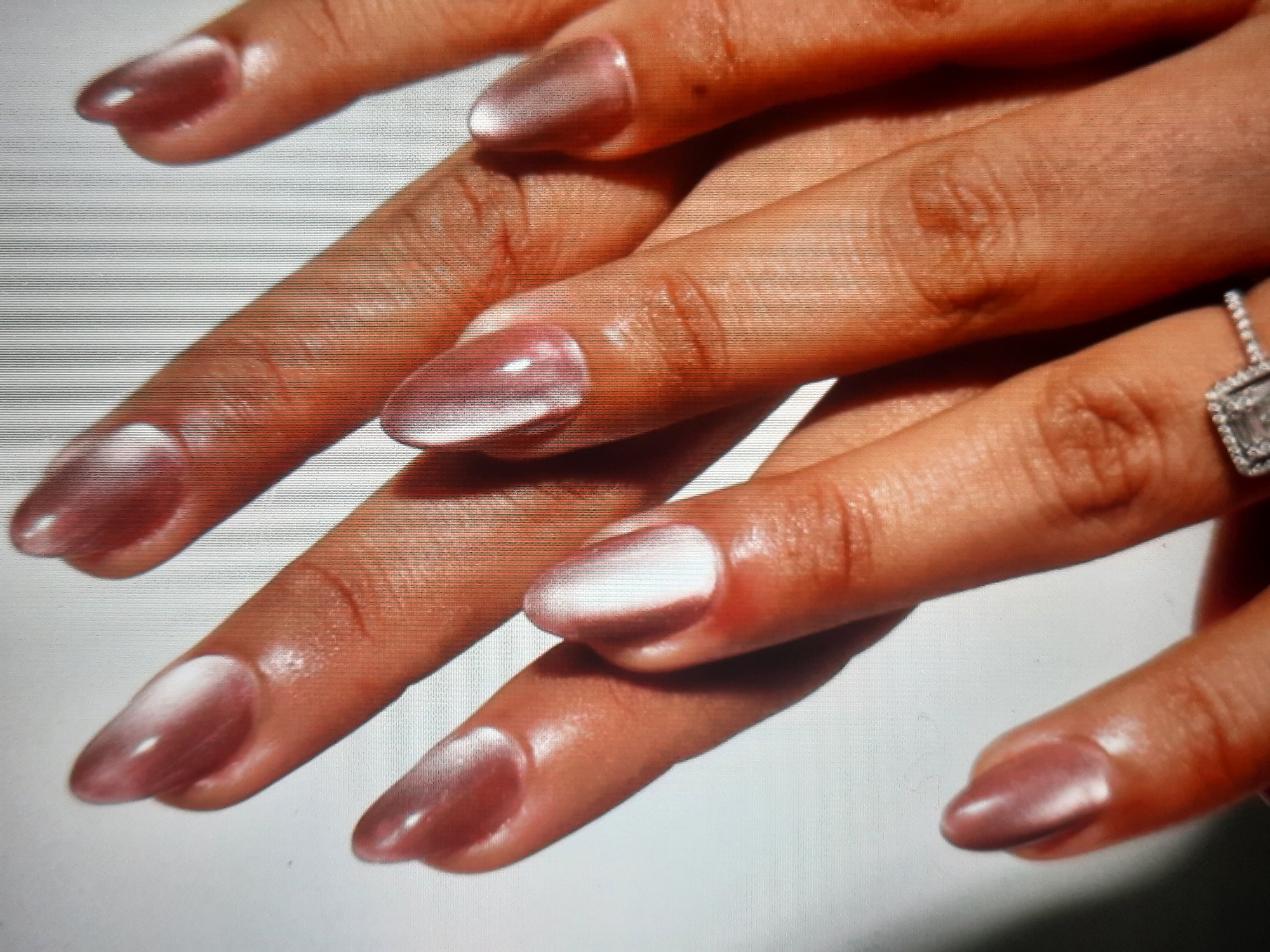 The image is a close-up, overhead shot of a person's hands, featuring manicured fingernails with a pearlescent, translucent pink finish. The nails are almond-shaped and exhibit a cat-eye effect, with a bright streak of light running through each one. The hands are positioned in a way that showcases the nails, with fingers overlapping and slightly curled. A portion of a silver ring with square-cut stones is visible on the ring finger of the lower hand. The background is a plain white surface, suggesting a studio or controlled lighting environment. The skin tone appears to be warm. There is no information about the location in Rosenheim, Germany, as this is a close-up of hands and does not contain any identifiable landmarks or context related to a specific geographical location.
