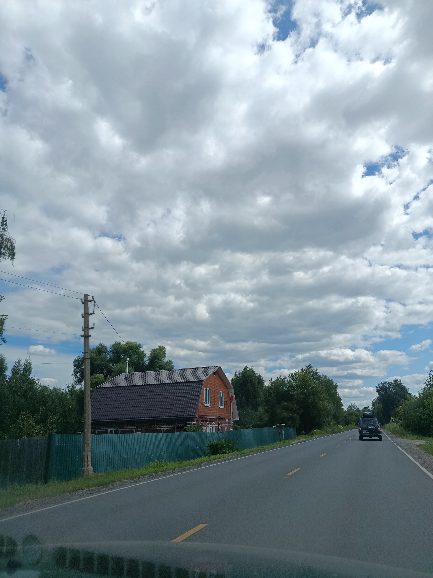 A roadside scene in Снопок Старый, Russia, on a cloudy day. A two-story wooden house with a dark-tiled roof is visible on the left, behind a green corrugated metal fence. A utility pole with multiple insulators stands in front of the house. The road is asphalt with yellow lane markers and stretches into the distance. A black SUV with a roof rack is driving away on the right side of the road. Trees and vegetation line the sides of the road. The sky is filled with dramatic white and gray clouds.