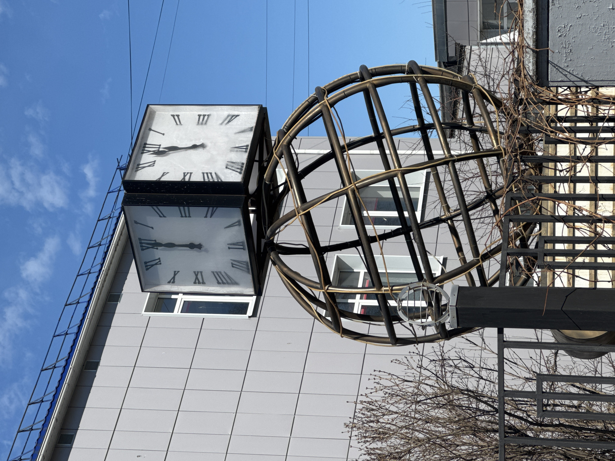 A low-angle shot depicts a city clock in Chaikovsky, Russia. The clock is square, double-sided, and black-framed, with white faces and Roman numerals. The clock hands on both sides indicate a time around 9:15. Beside the clock is a spherical cage-like structure made of metal bars. Behind the clock and sphere is a multi-story building with a light gray tiled facade and white-framed windows. Thin wires stretch across the blue sky above the scene. To the right of the sphere, vegetation, including bare vines and tree branches, is visible, along with a section of a dark gray metal grid-like structure. The image has been rotated to present the scene sideways.