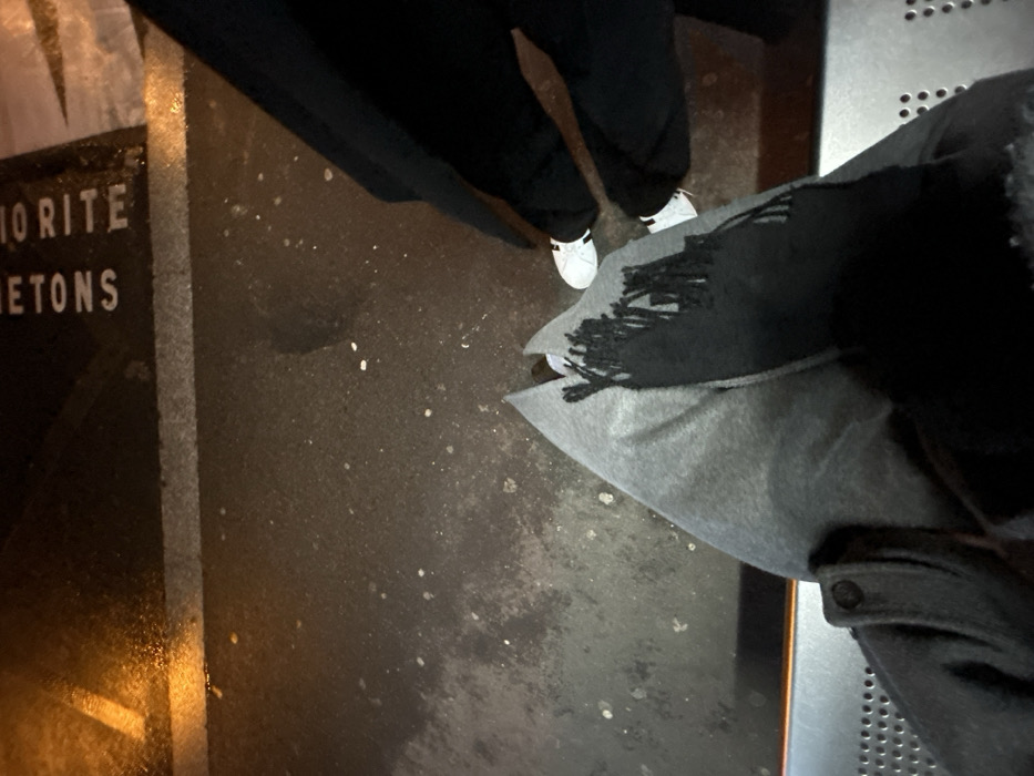 A top-down shot shows three sets of clothing on an outdoor surface. Two sets of legs are clad in black pants, with one person wearing white shoes. A gray and black scarf and jacket are visible in the foreground. In the background, part of a sign is seen, bearing the words "IO RITE" and "ETONS". The ground is dark and textured, showing signs of moisture. A metal grate is visible on the right side of the frame.