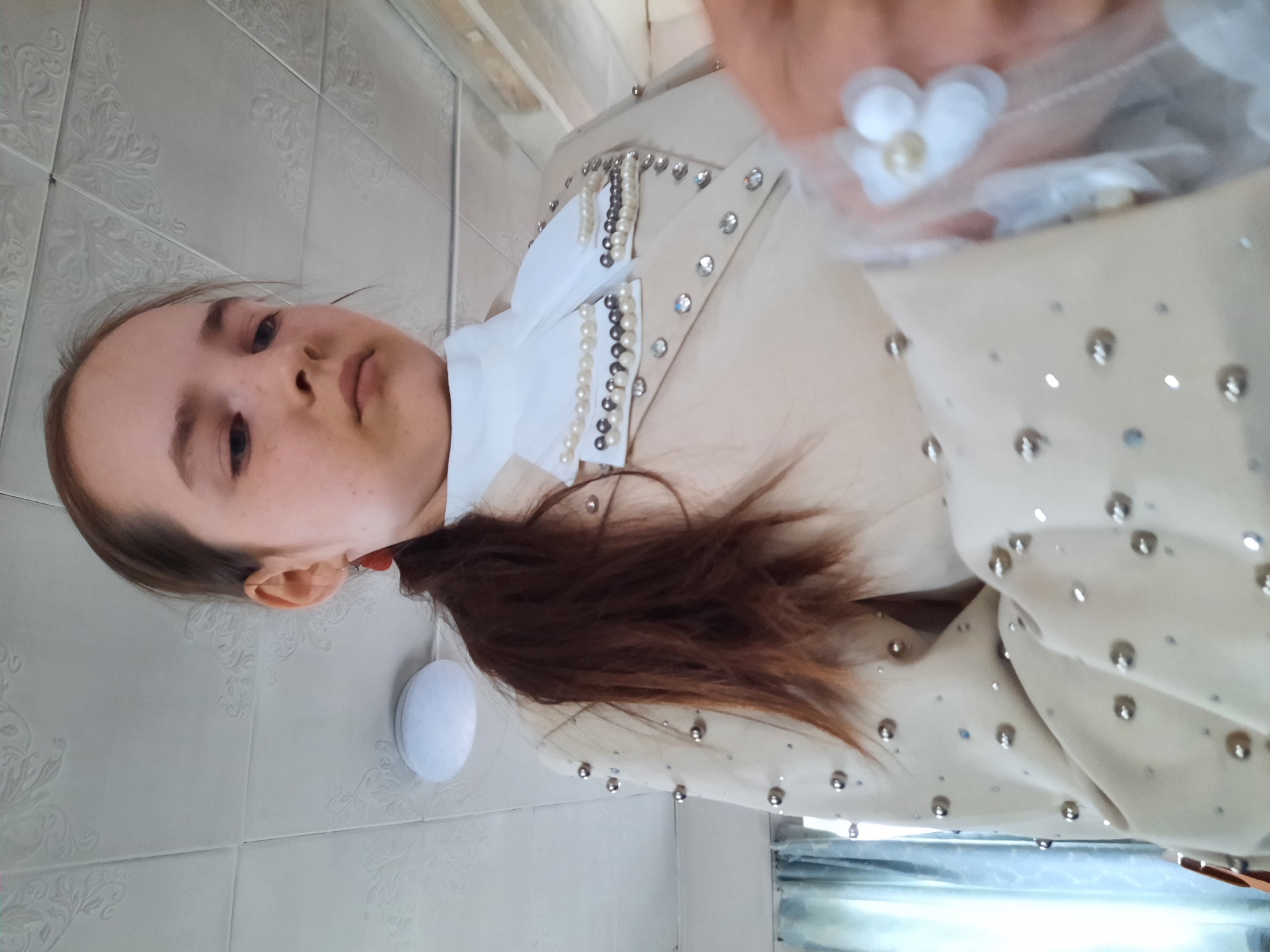 A person with brown hair, gathered in a ponytail, is wearing a light-colored garment adorned with silver studs and pearls, with a white collar. The person is positioned against a tiled wall. The location is unknown.
