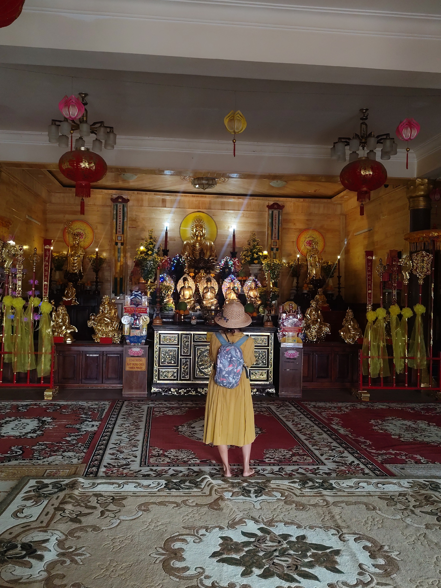 In the city of Lạc Dương, Vietnam, a woman stands facing an altar in a temple, wearing a straw hat and a yellow dress. She has a backpack on her shoulders and is barefoot. The altar is adorned with numerous golden statues of deities, flowers, and offerings. Red lanterns hang from the ceiling, and the floor is covered with ornate carpets. The walls are decorated with wooden panels and religious symbols. The scene suggests a spiritual or cultural site, possibly a Buddhist temple.