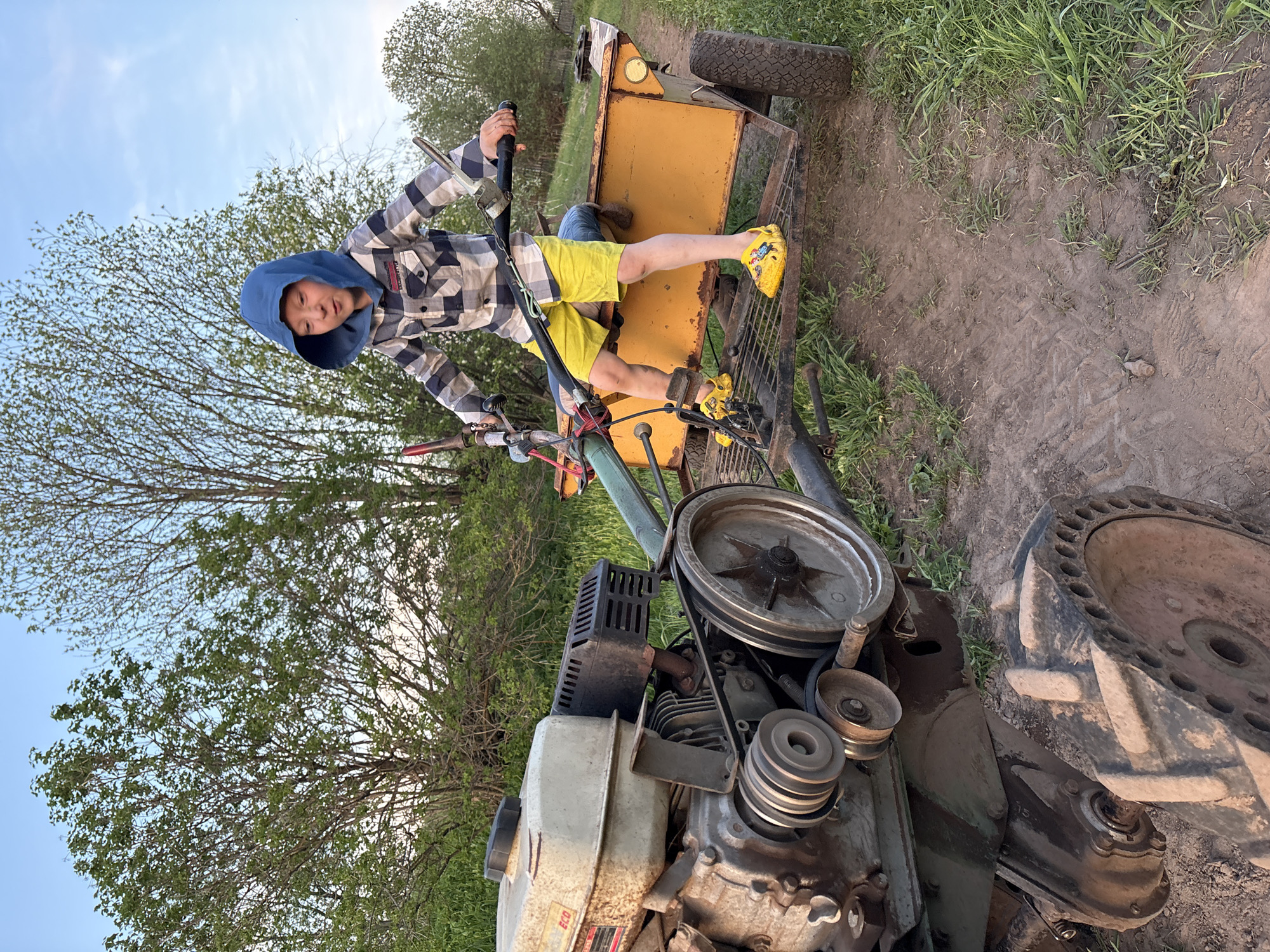 A young person wearing a checkered hooded shirt and yellow shorts is sitting on a yellow and green tractor-like vehicle with a trailer. They are wearing yellow footwear. The vehicle is located on a dirt surface with patches of grass. Trees are visible in the background, and the sky is partially visible. This image was taken in Nizhnyaya Chermoda, Russia.