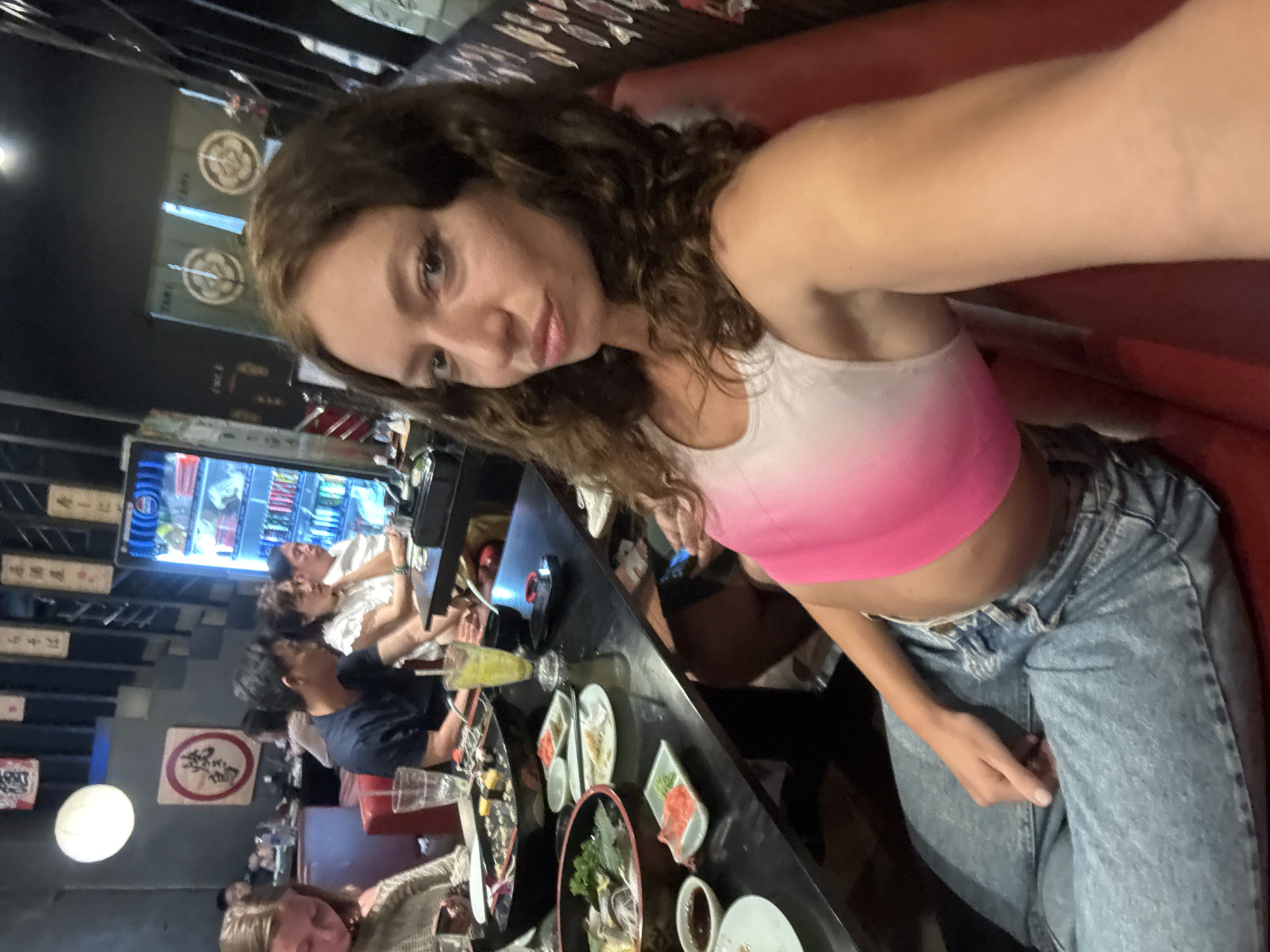 A woman with curly brown hair is taking a selfie in what appears to be a restaurant or bar in Đà Nẵng, Vietnam. She is wearing a white and pink crop top and blue jeans. In the background, other people are visible seated at the counter, and plates of sushi and other dishes are on the table. There is Vietnamese signage and a drinks fridge in the background, and a globe-shaped light fixture. The scene appears casual and social.