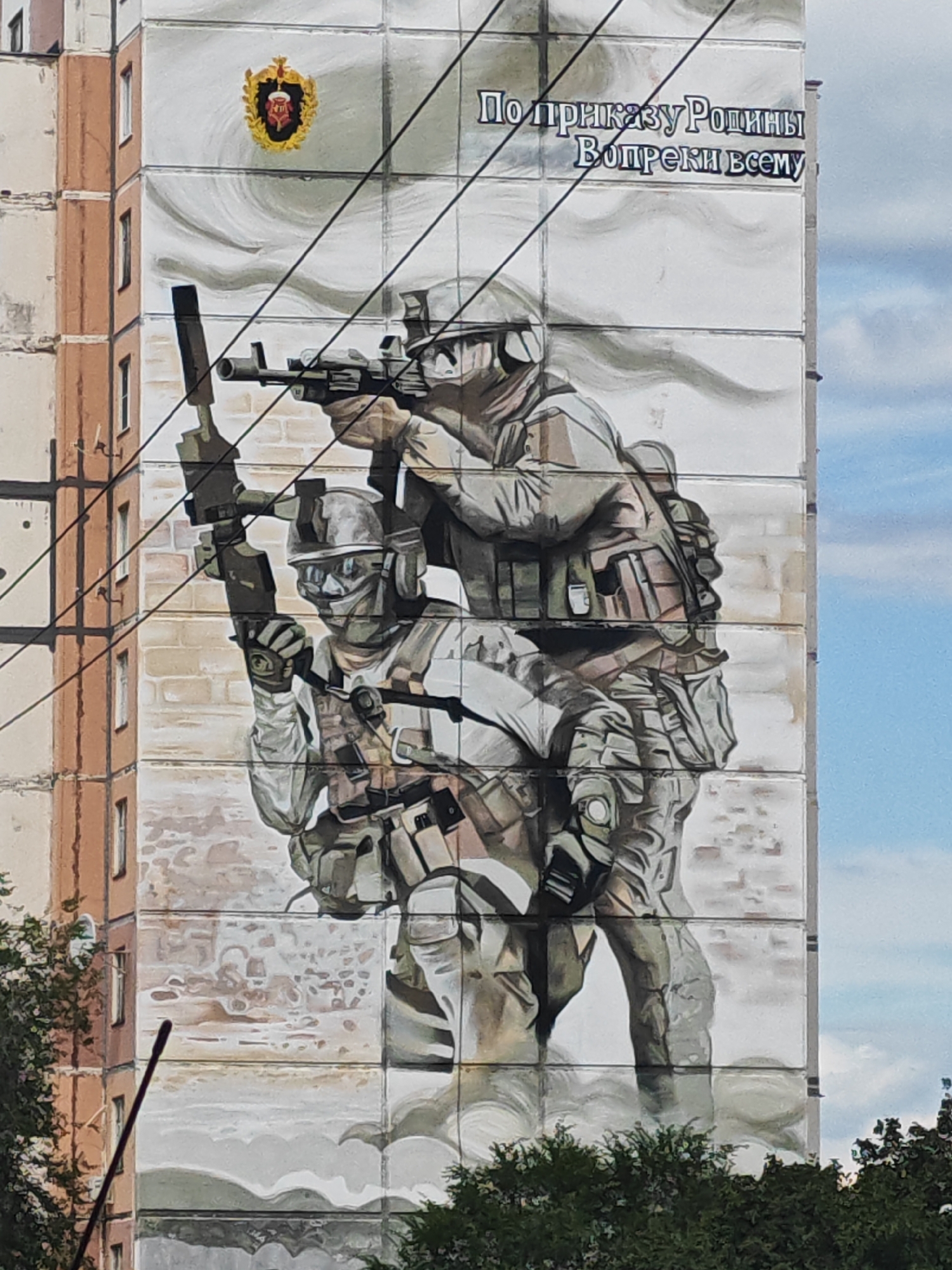 The image shows a mural painted on the side of a multi-story building in Tolyatti, Russia. The mural depicts two soldiers in camouflage uniforms and tactical gear, carrying rifles and aiming them. The soldier in the foreground is kneeling, while the soldier behind him is standing. Above the soldiers, there is a crest with a red and gold emblem and text in Russian that reads "По приказу Родины Вопреки всему" (By order of the Motherland, against all odds). The mural is painted on a section of the building's facade that has vertical lines, possibly indicating the seams between prefabricated concrete panels. The building itself appears to be a typical Soviet-era apartment block, with visible windows and balconies. The mural is set against a backdrop of a cloudy sky. Trees and other foliage are visible at the bottom of the image, suggesting an outdoor urban environment. The mural's presence on the building indicates it is part of the urban landscape of Tolyatti.
