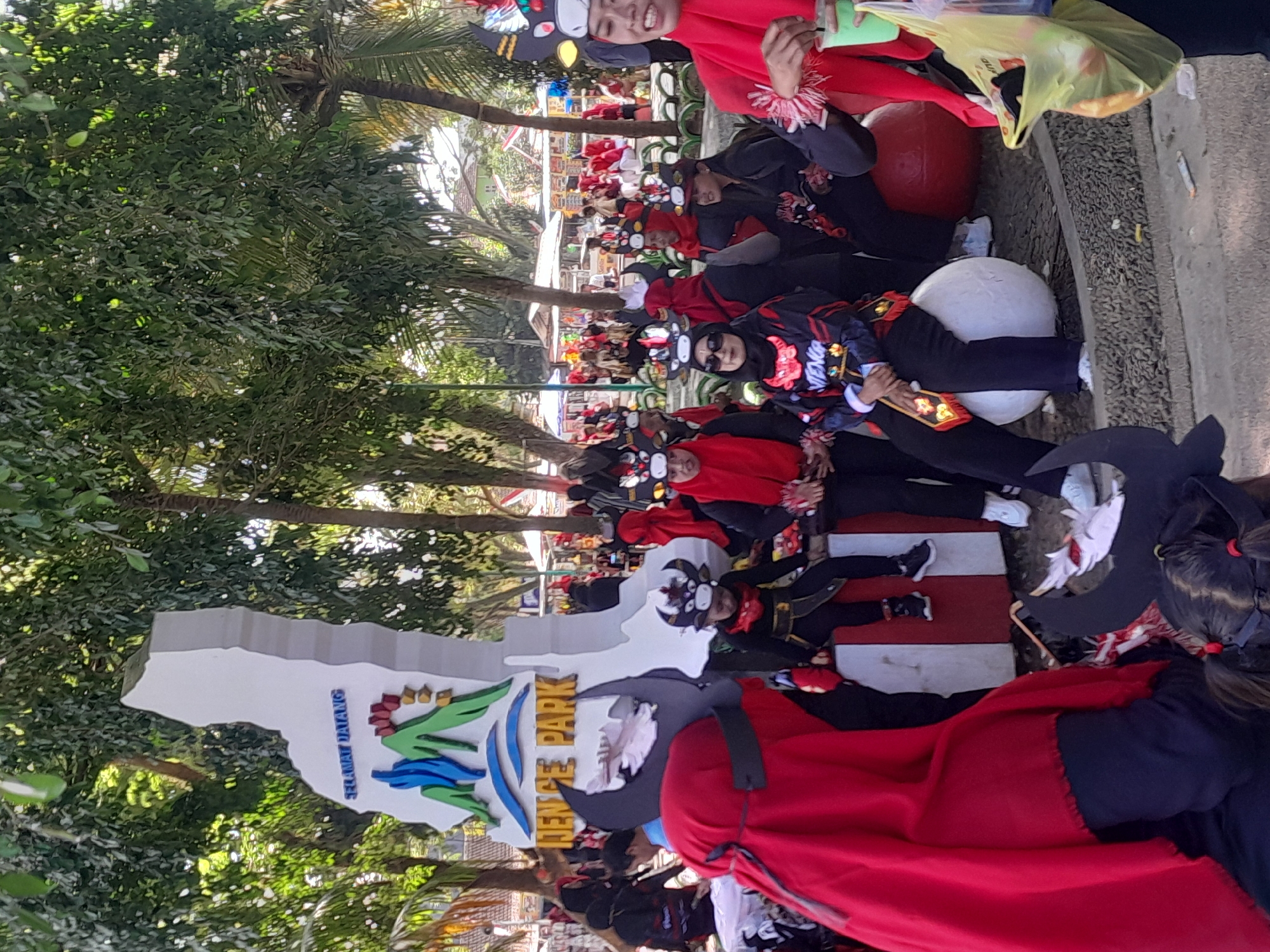 A group of people are gathered in front of a sign that reads "SELAMAT DATANG" (Welcome) and "JENC E PARK" in Genteng Kulon, Indonesia. The people are dressed in red and black costumes with bull-like headpieces. Some are seated on large white and red balls. In the background, there are trees and tents that suggest a park or outdoor event. One person on the right is holding a green cup and a yellow plastic bag.
