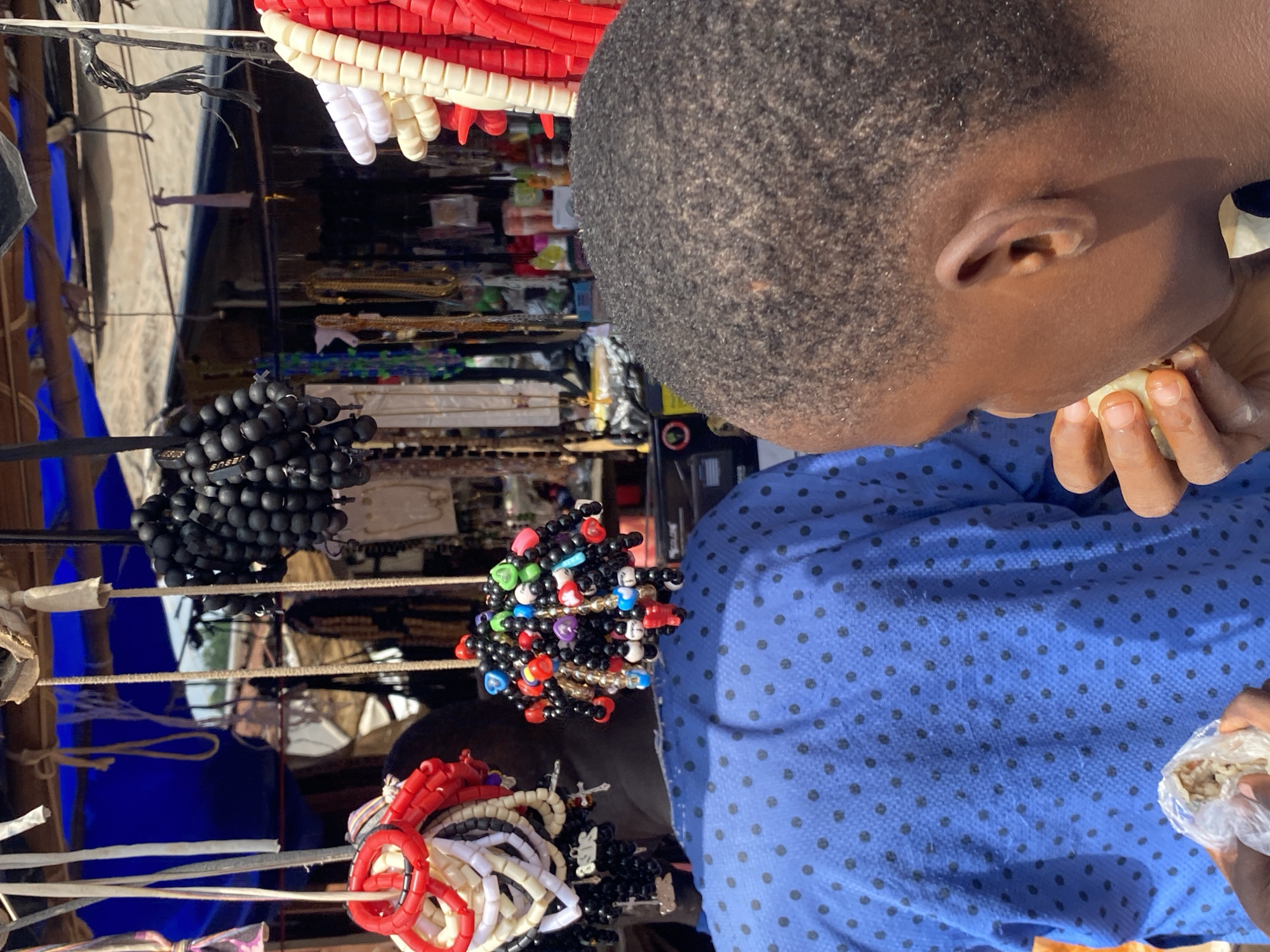 In Nguzu Edda, Nigeria, two individuals are at a market stall adorned with various beaded items. One person, seen from the back wearing a blue shirt with black polka dots, is partially obscured by their own shoulder and torso. In front of them, a second individual, a young male, is shown in profile from the right, facing left and eating. The market stall itself is set up under a makeshift roof, with a blue tarp visible in the background. Hanging from the stall are numerous strands of beads, including black, red, white, and multi-colored varieties. Some strands are clustered together, forming spherical shapes, while others hang in long strings. One cluster of black beads has a tag with the word "JESUS" printed on it. The overall scene depicts a bustling market environment with an emphasis on the sale of beaded accessories.