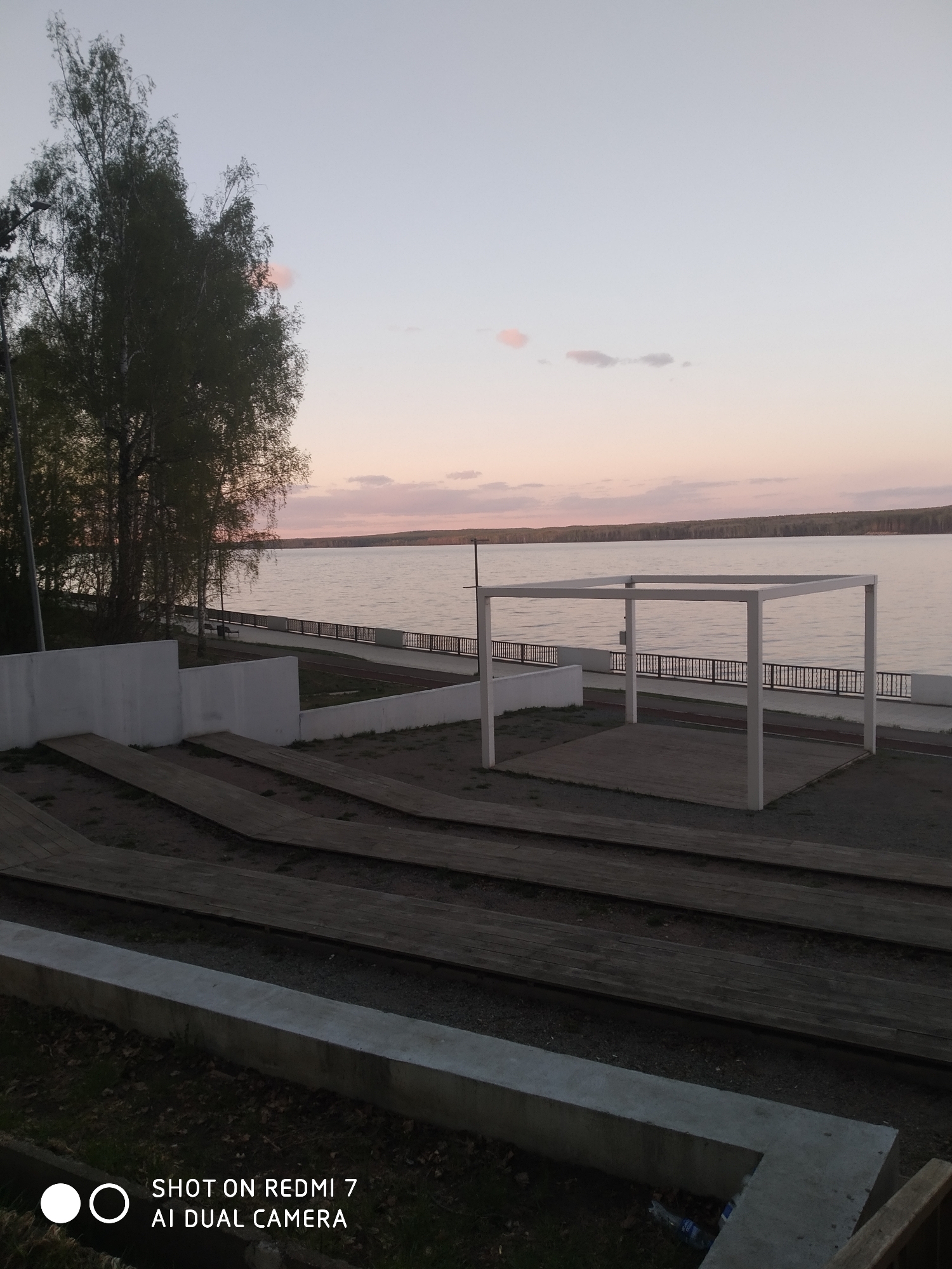 The image depicts a waterfront park scene in Chaikovsky, Russia. In the foreground, there's a tiered seating area with horizontal wooden planks supported by concrete risers. Moving towards the middle ground, there's a low white wall separating the seating area from a paved walkway along the water. A white square gazebo-like structure stands on a concrete base. Beyond the walkway, there is a railing and then a body of water, likely a river or lake. Across the water, there's a treeline. The sky is a soft gradient of pinks and blues, suggesting either sunrise or sunset. A few sparse trees are visible to the left of the seating area. The image was taken with a Redmi 7 AI Dual Camera phone, judging from the watermark at the bottom.