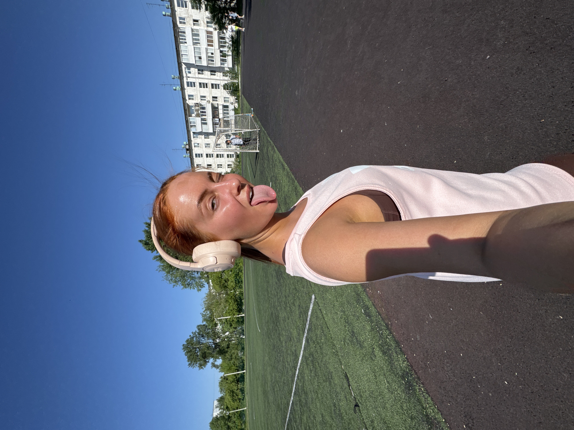 A young woman with reddish hair is taking a selfie on a sunny day. She is wearing headphones and a light pink tank top. Her tongue is sticking out. The photo appears to be taken next to a sports field with a running track and a soccer field. In the background is a multi-story building with many windows. Based on the style of architecture, the location is likely in Chaikovsky, Russia.