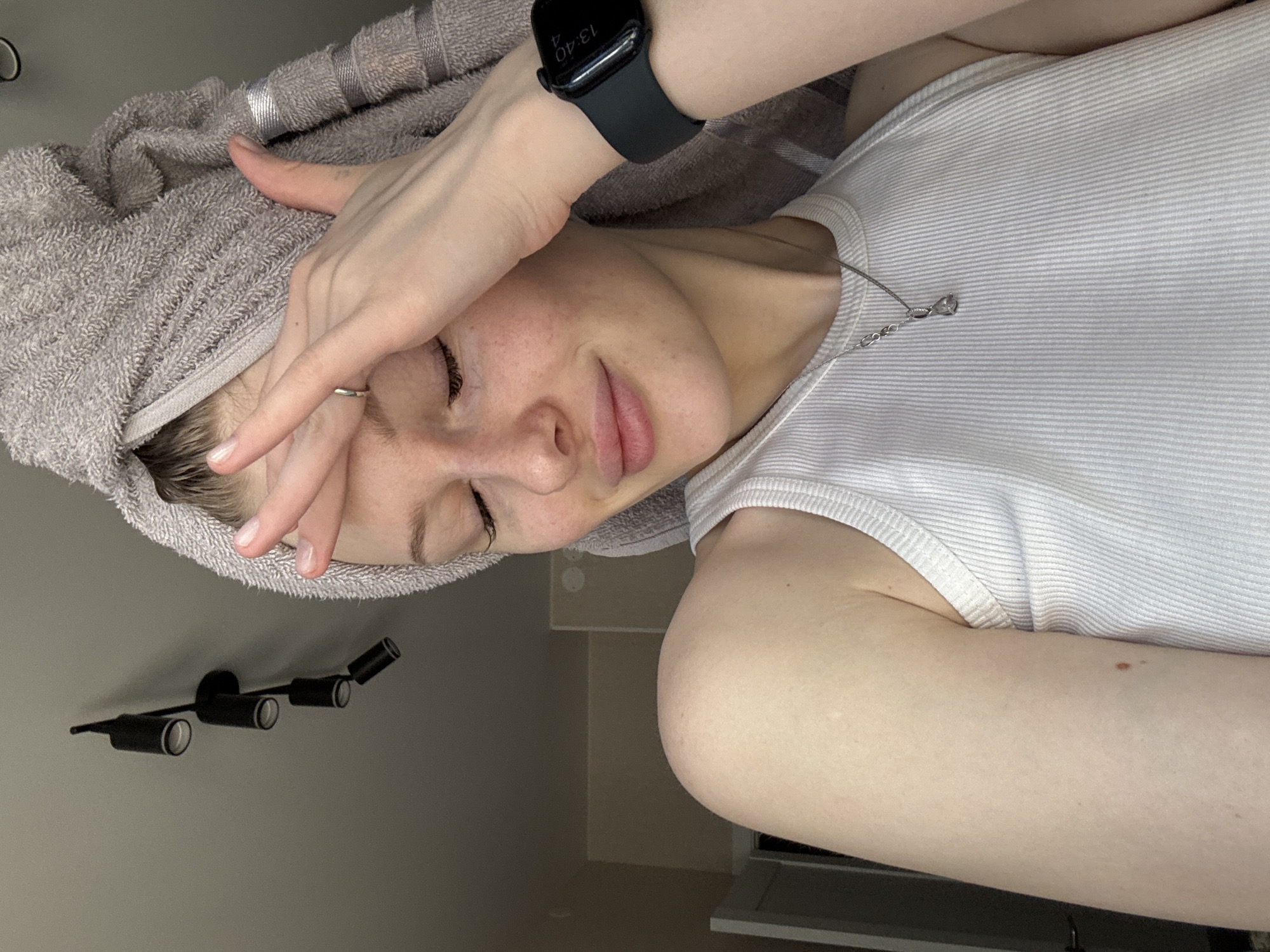 A woman is depicted in a close-up shot, seemingly indoors. She is wearing a white ribbed tank top and a smart watch. Her head is wrapped in a gray towel, and she is touching her forehead with her hand. She is wearing a necklace. The background reveals a wall with a black four-bulb lighting fixture. The location is unspecified, so it cannot be placed in Perm, Russia.