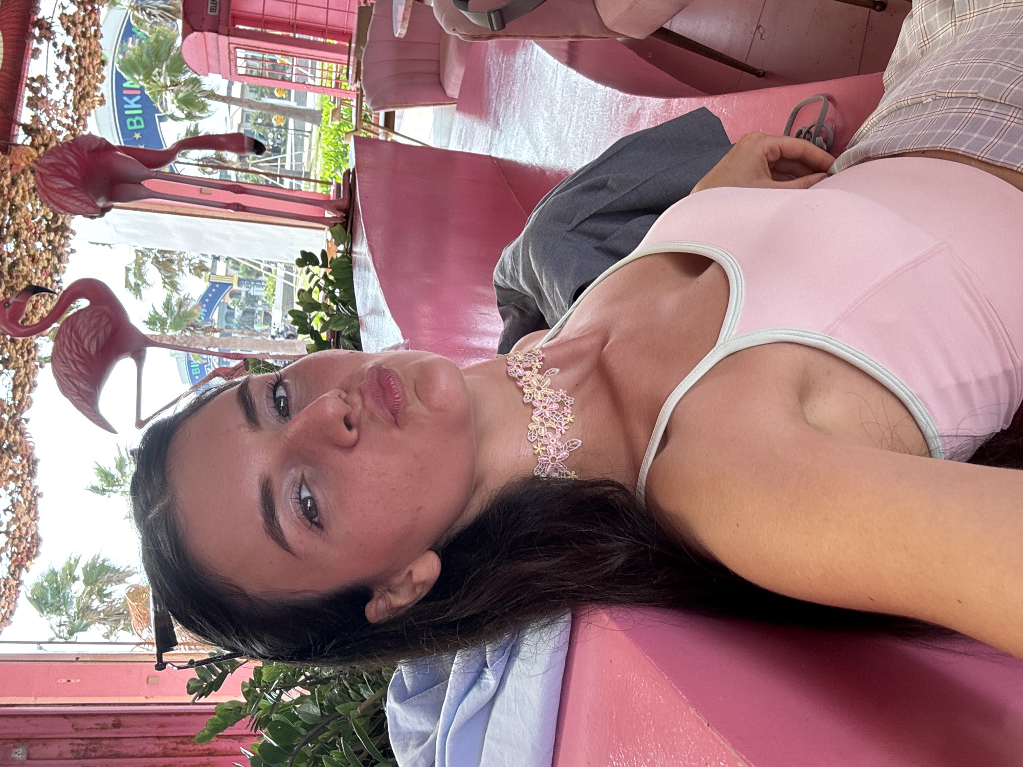A young woman with dark hair is seated in a pink booth, leaning against a pink surface. She is wearing a pink tank top with a light blue trim and a plaid skirt. Around her neck, she has a delicate, floral-patterned choker. She is looking at the camera with her lips puckered. In the background, pink flamingo decorations hang from the ceiling, and a bright pink phone booth is visible. Outside the window, palm trees and buildings suggest an outdoor setting. The scene appears to be a cafe or a similar establishment in Tien Thanh Ward, Vietnam, characterized by its predominantly pink decor.