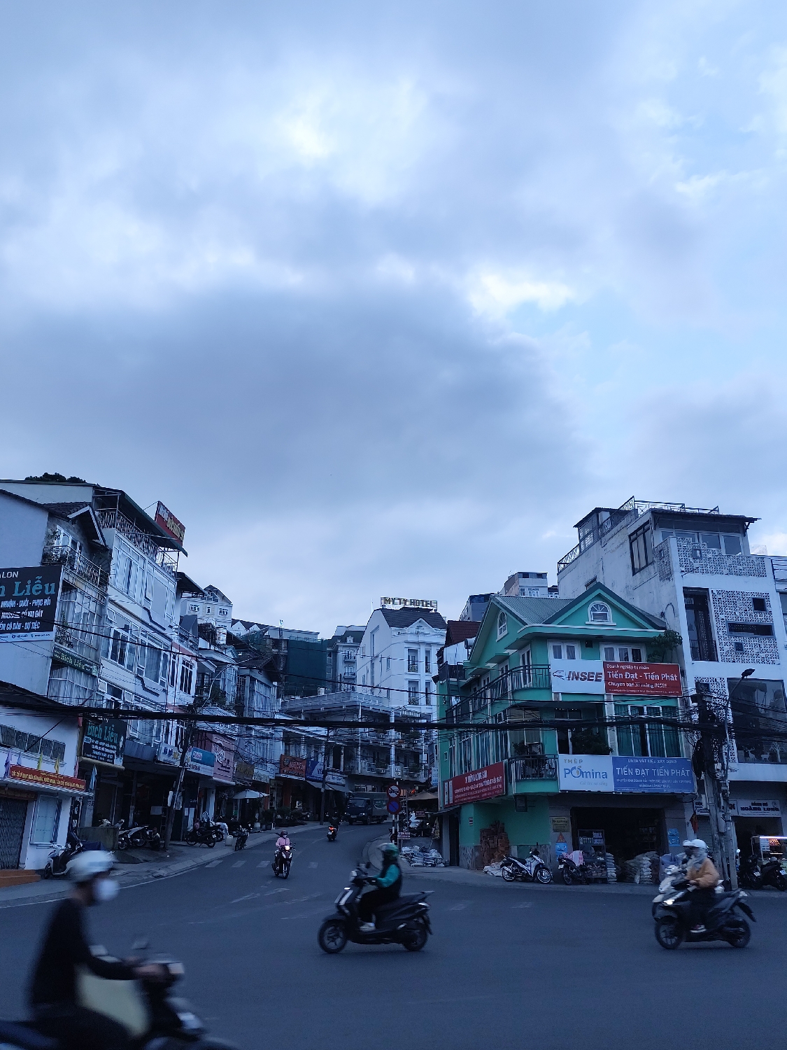 This image depicts a bustling street scene in Da Lat, Vietnam, under an overcast sky that suggests either early morning or late afternoon with diffused light. The central focus is a gently curving urban road lined with closely-packed, multi-story buildings that exhibit a blend of architectural styles, many featuring balconies and storefronts. A dense web of electrical wires is visible overhead, spanning across the street between buildings.
Numerous motorbikes are present, some parked along the roadside, others in motion with riders wearing helmets, and some also wearing face masks. One rider in the foreground is significantly motion-blurred. On the right side of the street, a prominent teal-green building stands out, beneath which construction materials, including bags of cement and rebar, are stacked outside a store.
Visible text on various signs confirms the Vietnamese location and indicates commercial activity. Notable signs include "MY TY HOTEL," "INSEE Tiến Đạt - Tiến Phát" which advertises "Chuyên bán Xi măng INSEE" (specializing in selling INSEE Cement), and "THÉP Pômina VẬT LIỆU XÂY DỰNG" (Pômina Steel Construction Materials). Other fragmented Vietnamese text includes "Liễu," "NHUẬN - DUỜI - PHỤC HỒI," and "ÊM CÔ DÂU - DỰ TIỆC." The scene captures everyday urban life, characterized by active traffic and ongoing commercial operations in a city with subtly hilly terrain.