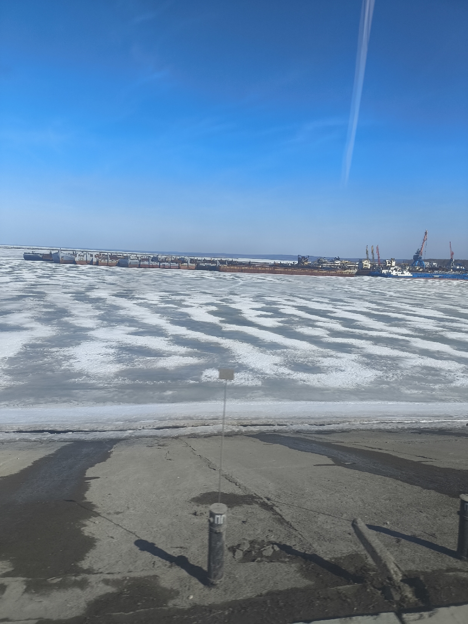 The image depicts a frozen body of water with a ship or barge docked at the harbor, presumably in the city of Chaikovsky, Russia. In the foreground is a paved area. The sky is blue with a contrail visible. The water is partially covered with ice. The ship or barge is long and appears to be docked with several cranes visible in the background. No people are visible in the image.