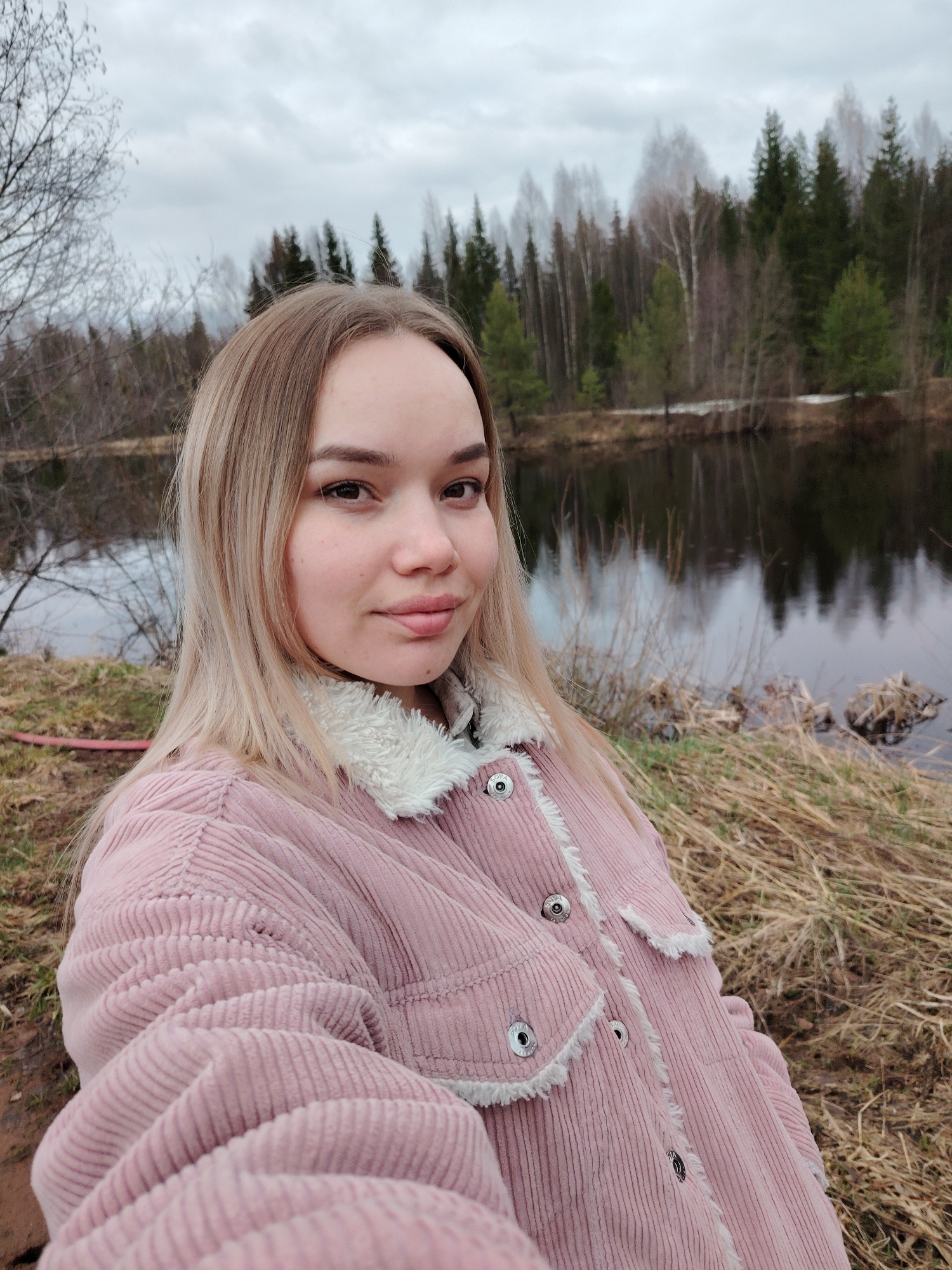 A young woman with blonde hair is captured in a selfie, posing outdoors next to a body of water, likely a river or lake, in what appears to be Chaikovsky, Russia. She is wearing a pink corduroy jacket with white fleece lining and silver buttons, which suggests cool weather. Her expression is neutral with a slight smile as she looks directly at the camera.
The background features a tranquil natural setting. On the opposite bank of the water, a dense forest stands, comprising both coniferous and deciduous trees, some of which are bare, indicating late autumn, winter, or early spring. A patch of snow is visible on the far bank, further supporting this seasonal assessment. The sky is overcast and grey, suggesting a cloudy day. The immediate foreground shows dry, brown grass, and to the far left, a glimpse of a bare tree branch and a red hose or pipe is visible. The water reflects the trees and the sky, creating a muted, earthy color palette for the scene. There is no visible text in the image.