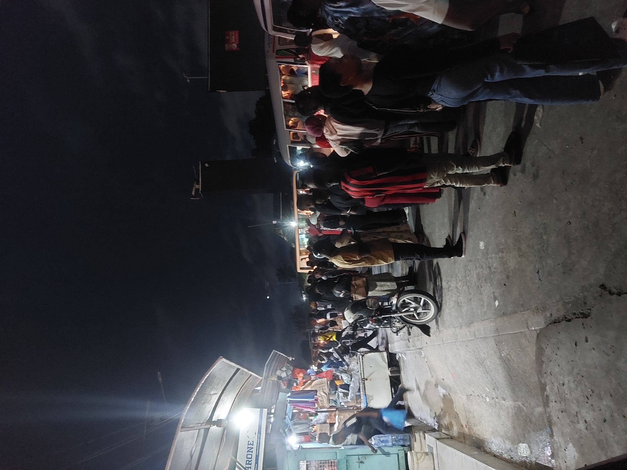 The image depicts a nighttime scene in Dar es-Salaam, Tanzania, likely a bustling street market or transport hub. Several people are boarding or queuing near a minibus, suggesting a public transport stop. Many individuals are dressed in casual clothing, including jeans and t-shirts. Some wear head coverings. A motorcycle stands parked nearby. The street features stalls with goods displayed under a canopy, indicating a marketplace. The environment appears to be urban, with artificial lighting illuminating the scene against the dark sky. The overall impression is one of urban activity and commerce in a developing city.