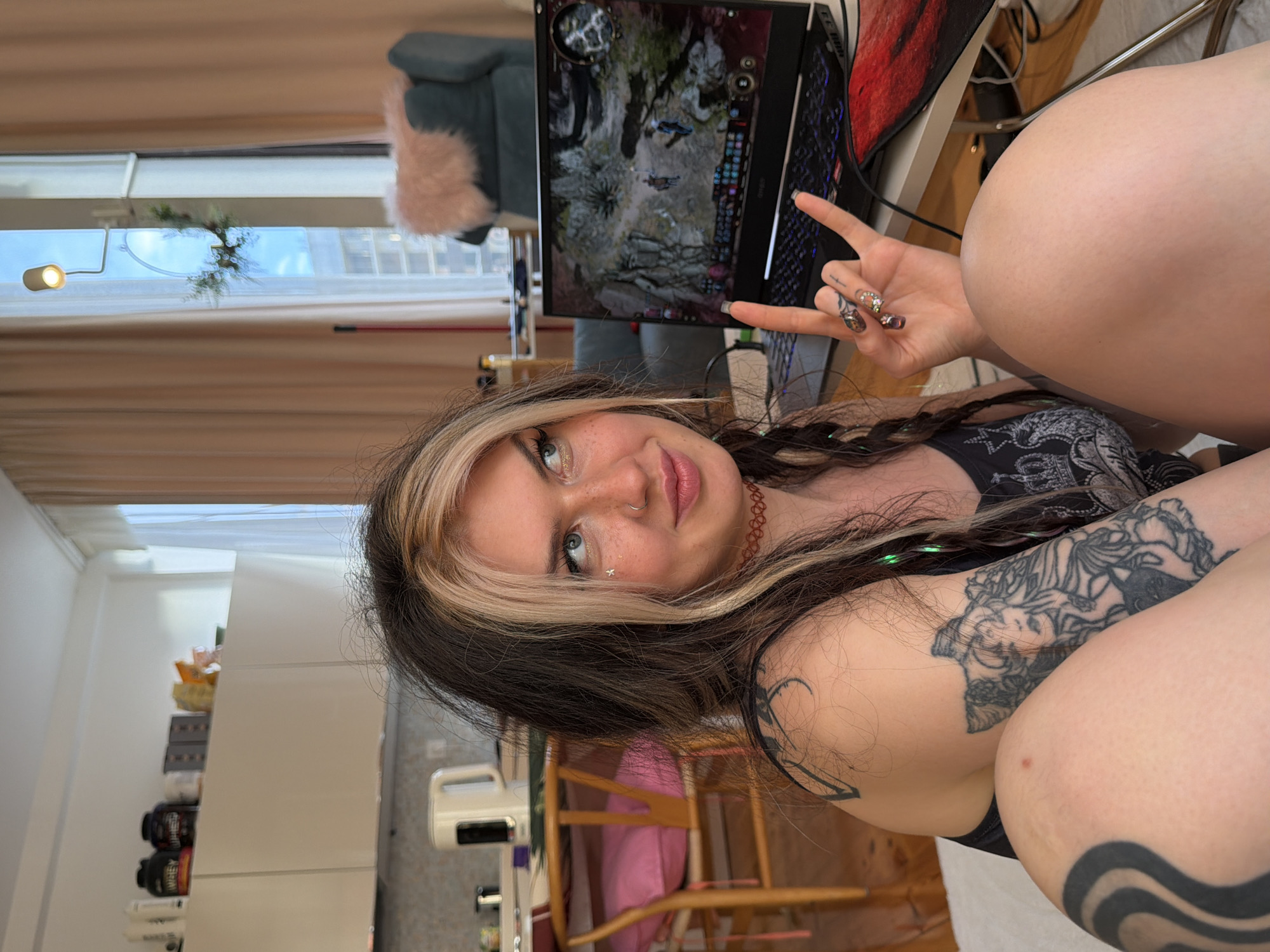 A young woman with long, dark brown hair with a blonde streak poses indoors, giving a peace sign with her left hand. She has tattoos on her arms and is wearing a black tank top with a design. A laptop is open in the background, displaying a video game screen. A blue chair with a pink fur cushion is partially visible behind the laptop. The room appears to be a residential space with a window and curtains. The woman is facing the camera. The location is in Budva, Montenegro.