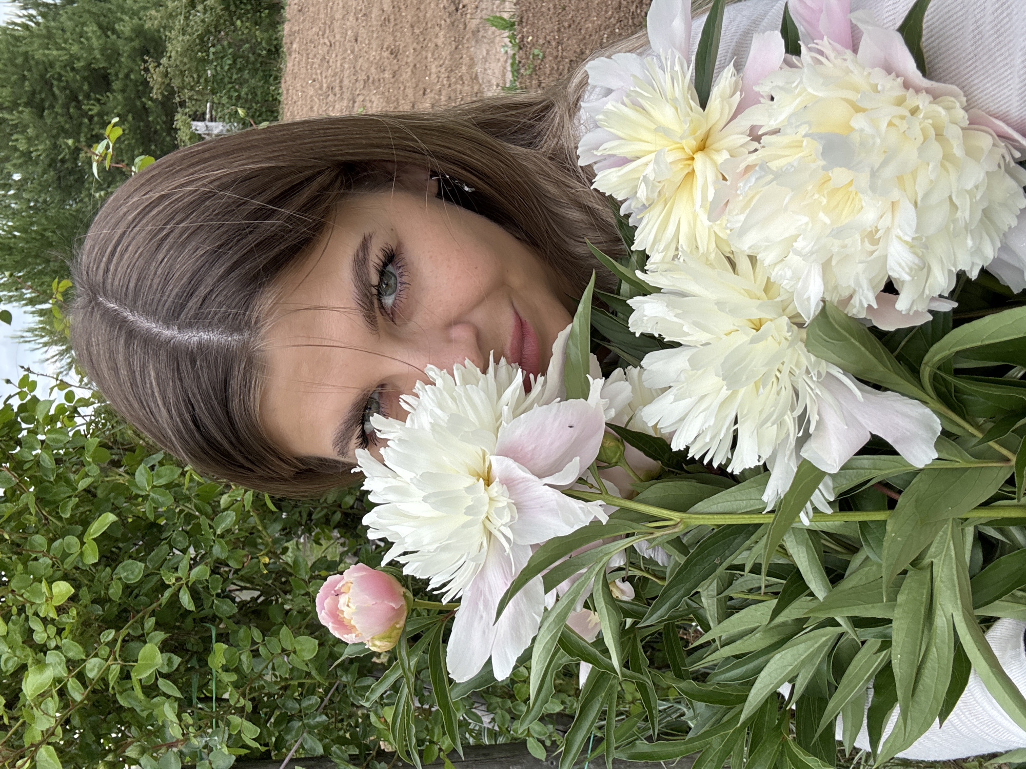 In the city of Fokino, Russia, a woman with long brown hair and green eyes is partially obscured by a bouquet of peonies. She is wearing a white top and is looking at the camera with a slight smile. The peonies are a mix of white and pale pink, with lush green leaves. The background features a garden with green foliage and a dirt path.