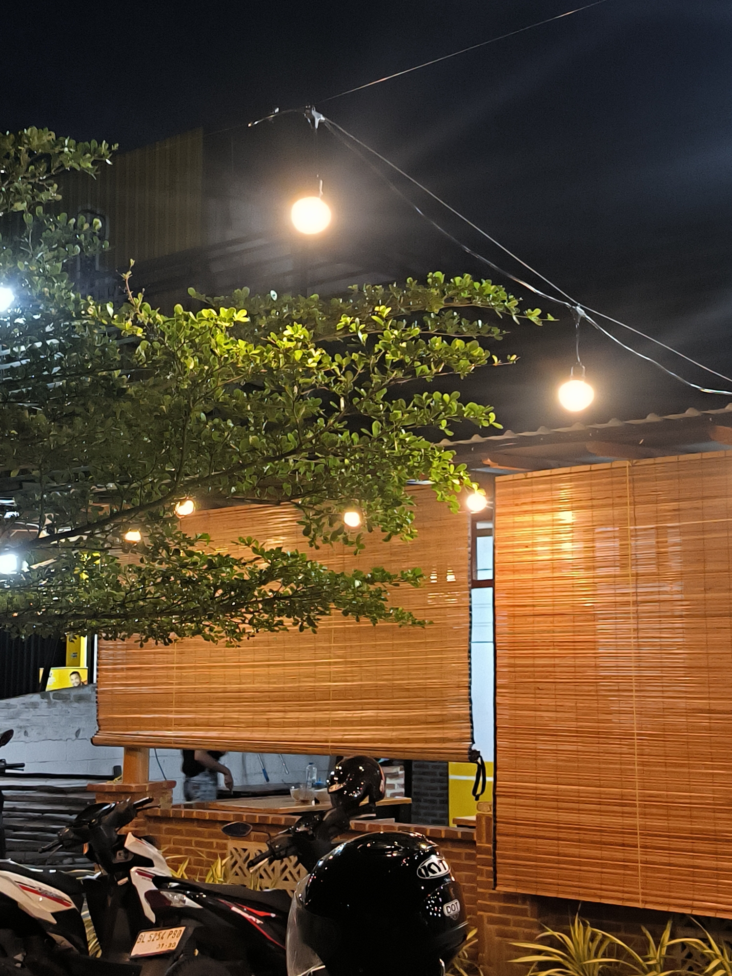 The image depicts an outdoor or semi-outdoor dining/gathering area at night, characterized by a warm and inviting ambiance. The dark night sky fills the upper portion of the frame, contrasting with the bright artificial lights.
A large, leafy green tree dominates the left side of the scene, its branches extending towards the center and intertwining with string lights. Several warm, glowing spherical light bulbs are strung across the scene, hanging from wires that stretch across the dark sky, providing illumination.
Below the tree and lights, multiple bamboo roll-up blinds serve as walls or coverings for openings, showcasing a natural, rustic aesthetic. A brick wall or base is visible beneath these blinds. In the background, a building or structure with horizontal paneling is faintly visible in the upper left.
The foreground is occupied by several parked motorcycles, with portions of their bodies and wheels visible. Two black motorcycle helmets are prominently placed on the bikes. One helmet clearly displays the brand "KYT" and the certification mark "DOT". A visible Indonesian license plate on one motorcycle reads "BL 5254 PBD" with "07•20" below it, indicating Aceh province (consistent with Banda Aceh).
Through an opening beneath one of the bamboo blinds, a person is faintly visible. Another person's face can be partially seen further in the background, behind some foliage and a yellow structure. The scene suggests a casual setting, likely an eatery or cafe, with patrons' vehicles parked nearby. Small, spiky plants are also visible at the bottom right, contributing to the outdoor atmosphere.