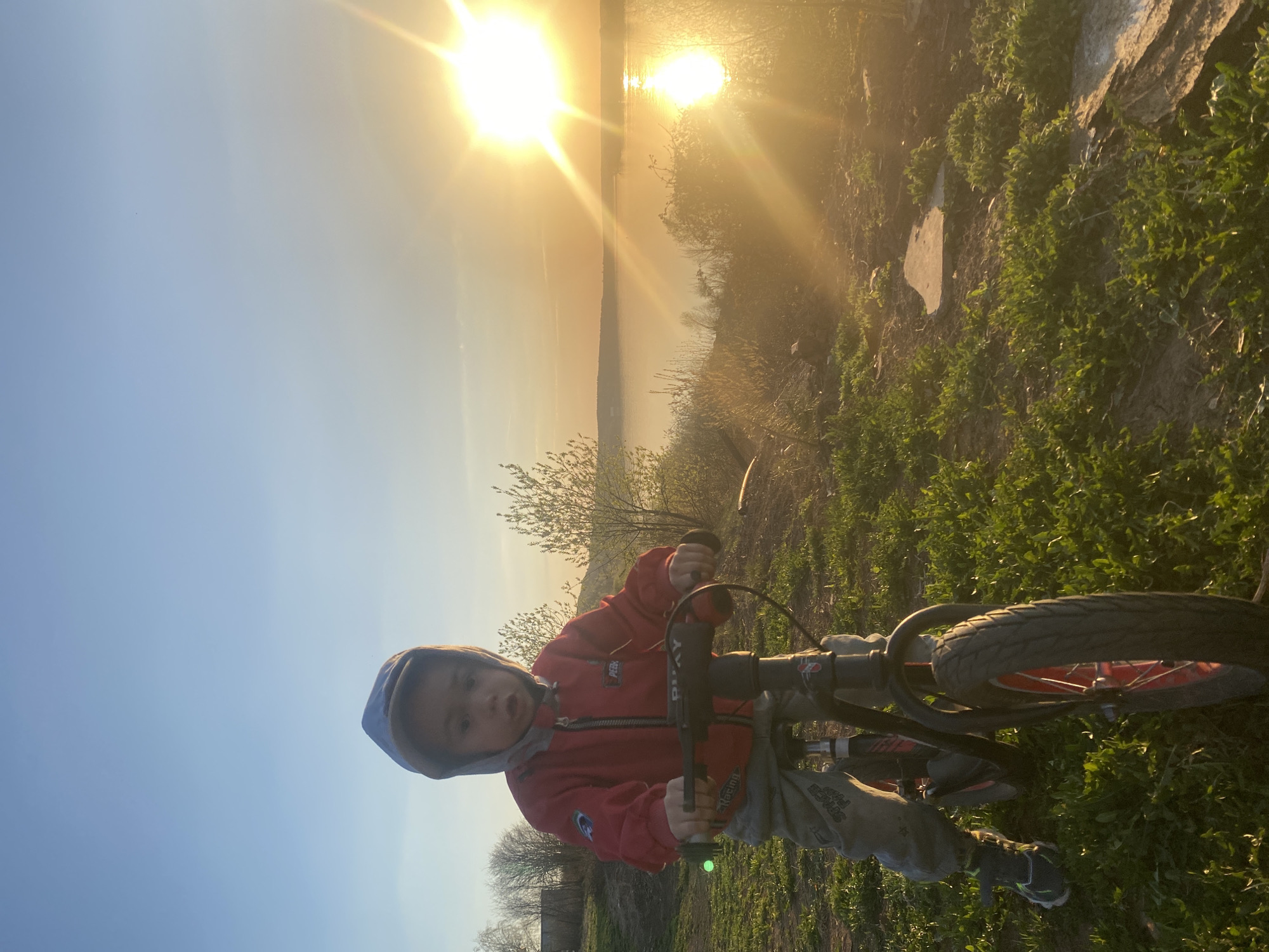 A young child in a red hooded jacket stands on a bicycle in a grassy area. The sun is setting or rising over a body of water in the background, creating a strong glare. The location is identified as Ольховка, Russia.